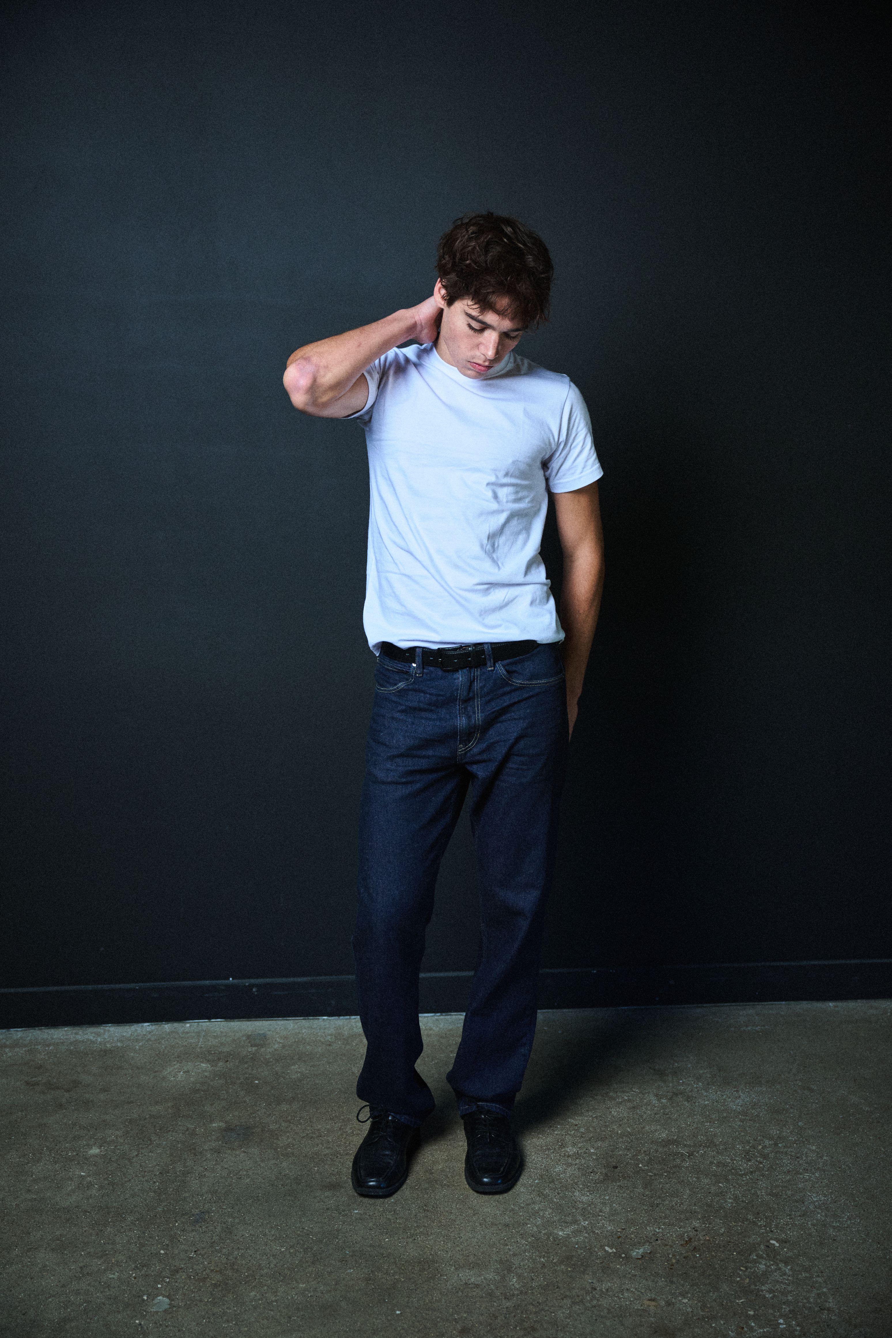 Male model in a white t-shirt and jeans leaning full length against a dark studio wall — Fashion & editorial photography, Austin A male model in a white t-shirt and jeans leaning full length against a dark studio wall.