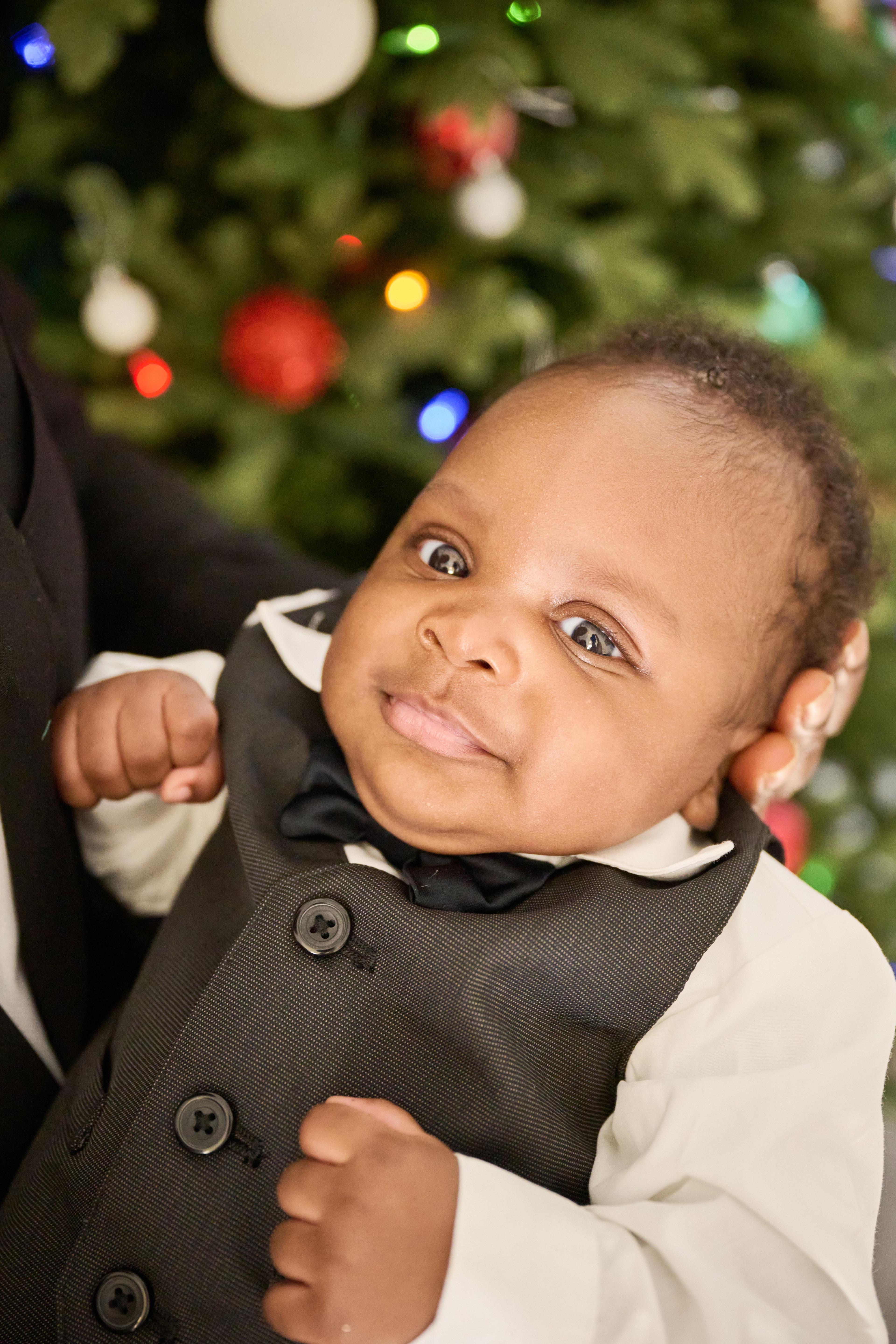 Baby in a waistcoat and bow tie smiling in front of a decorated tree — Seasonal Christmas & Fall photography, Austin A baby in a waistcoat and bow tie smiling in front of a decorated tree.