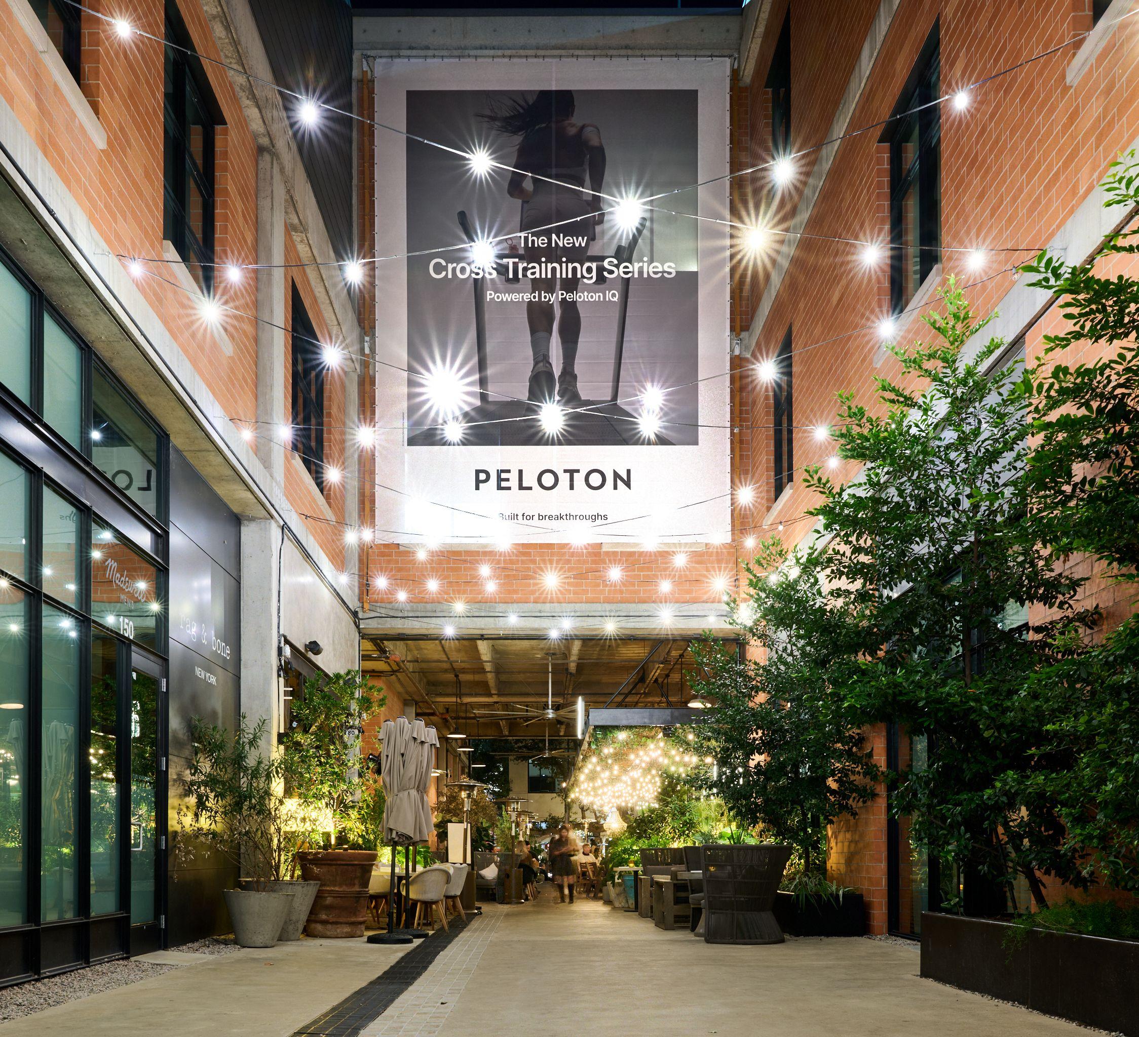 Peloton campaign banner hanging above a string-lit pedestrian alley at dusk — Outdoor media photography, Austin A Peloton campaign banner hanging above a string-lit pedestrian alley at dusk.