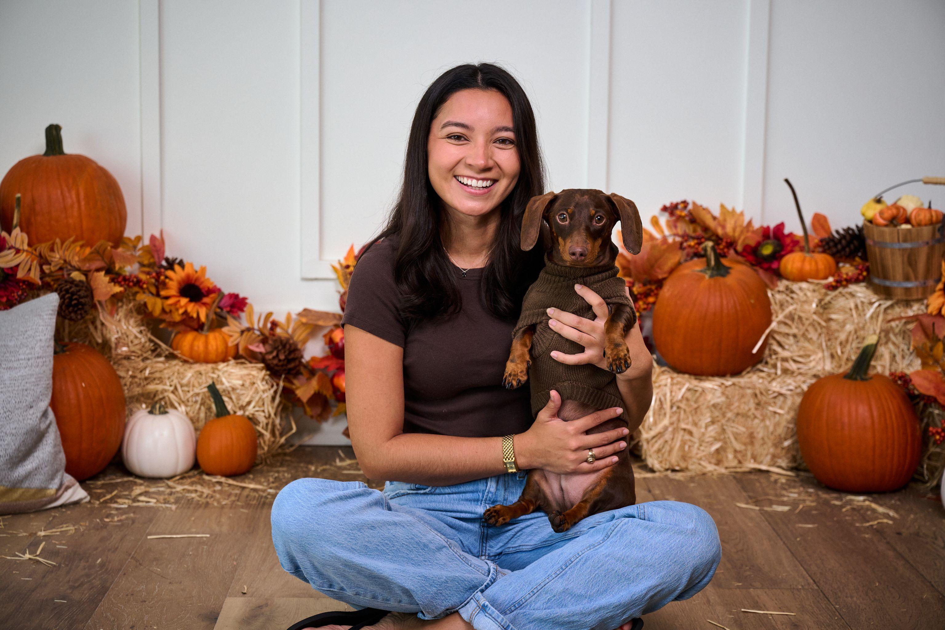 Young woman sitting cross-legged holding a dachshund on an autumn set with pumpkins — Mobile photo studio, Austin A young woman sitting cross-legged holding a dachshund on an autumn set with pumpkins and hay bales