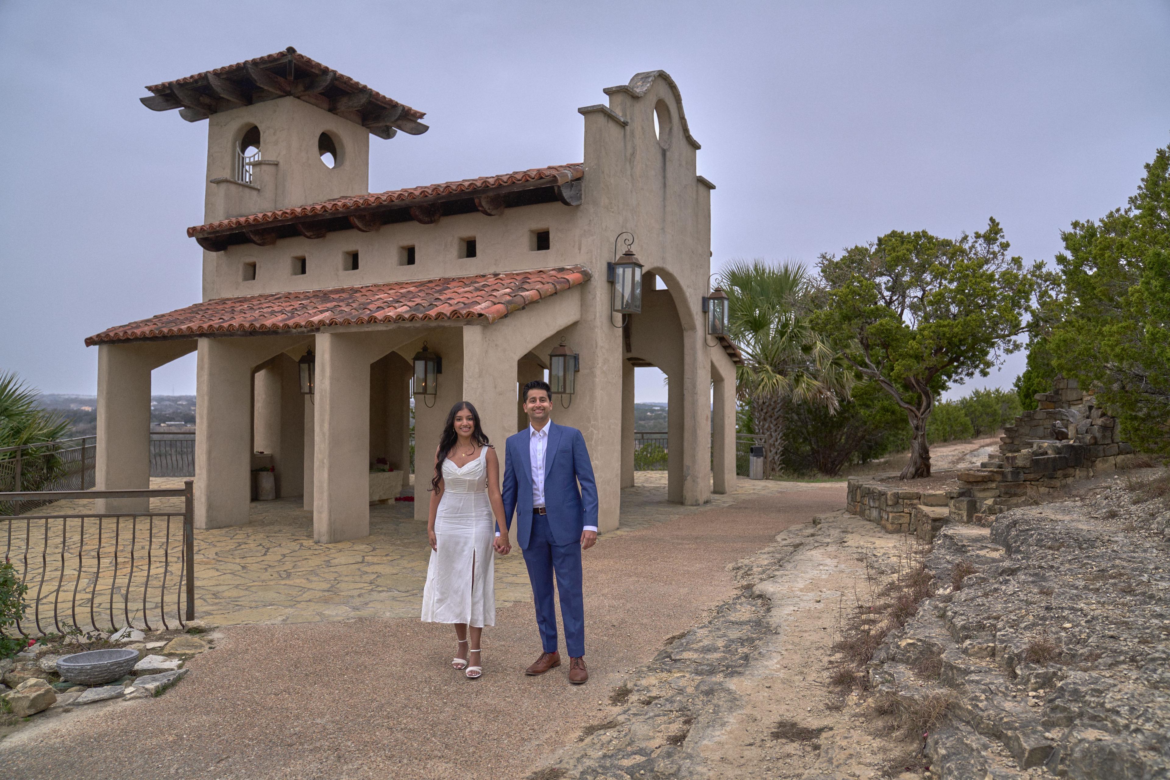 Couple standing small beneath the stucco arcade of Chapel Dulcinea, the red tile roof — Chapel Dulcinea photography, Austin A couple standing small beneath the stucco arcade of Chapel Dulcinea, the red tile roof and bell tower above them