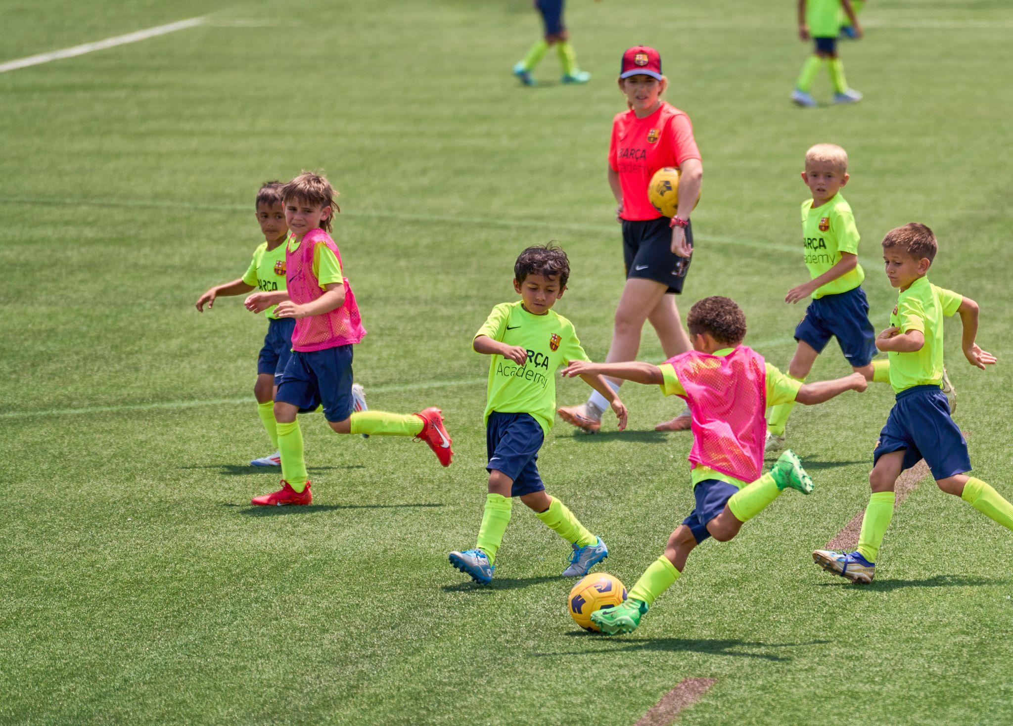 Young children in bright kits chasing a ball across a grass pitch with a coach — Sports photography, Austin Young children in bright kits chasing a ball across a grass pitch with a coach behind