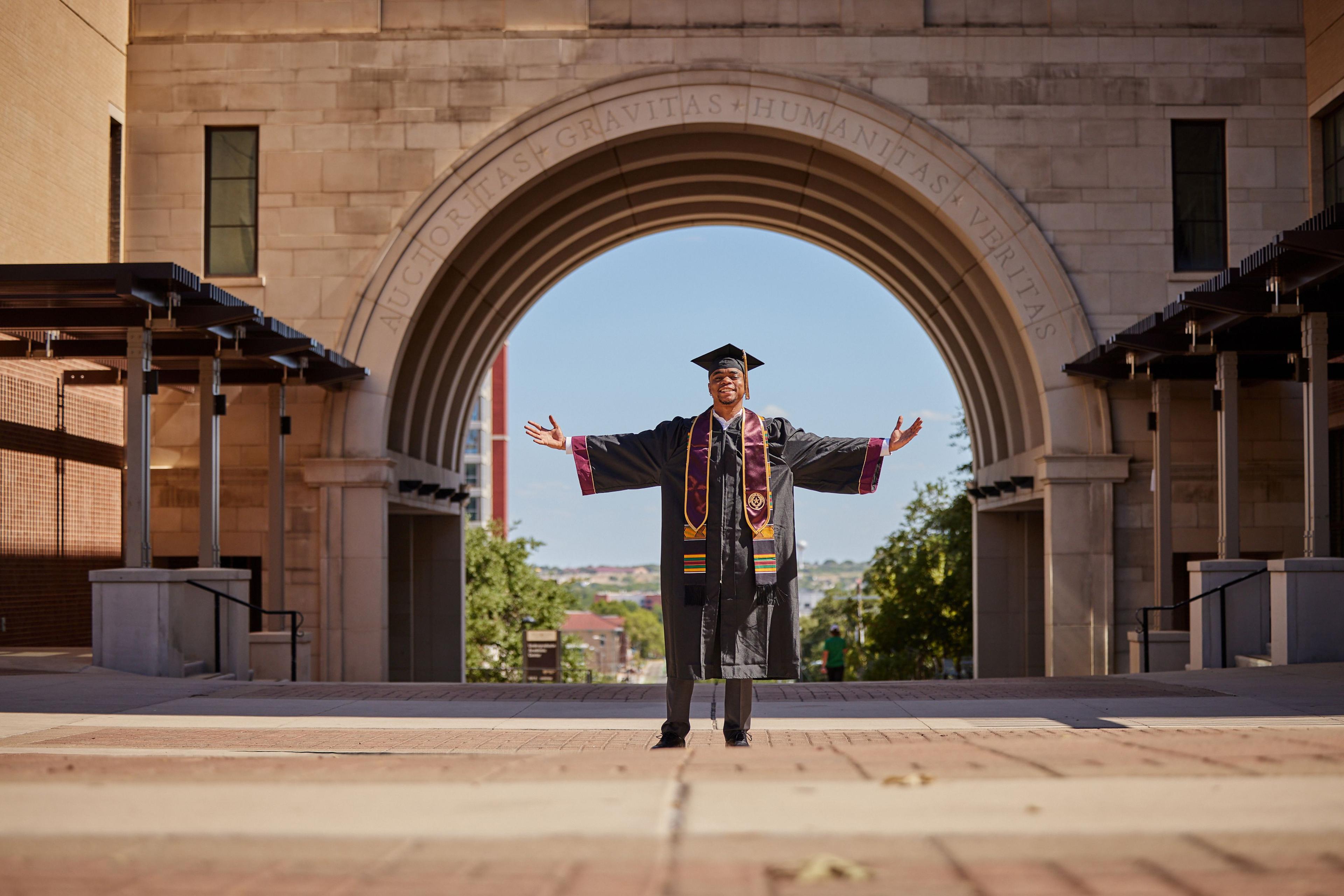 Graduate in cap and gown standing with arms outstretched inside the Texas State arch — Texas State University photography, Austin A graduate in cap and gown standing with arms outstretched inside the Texas State arch