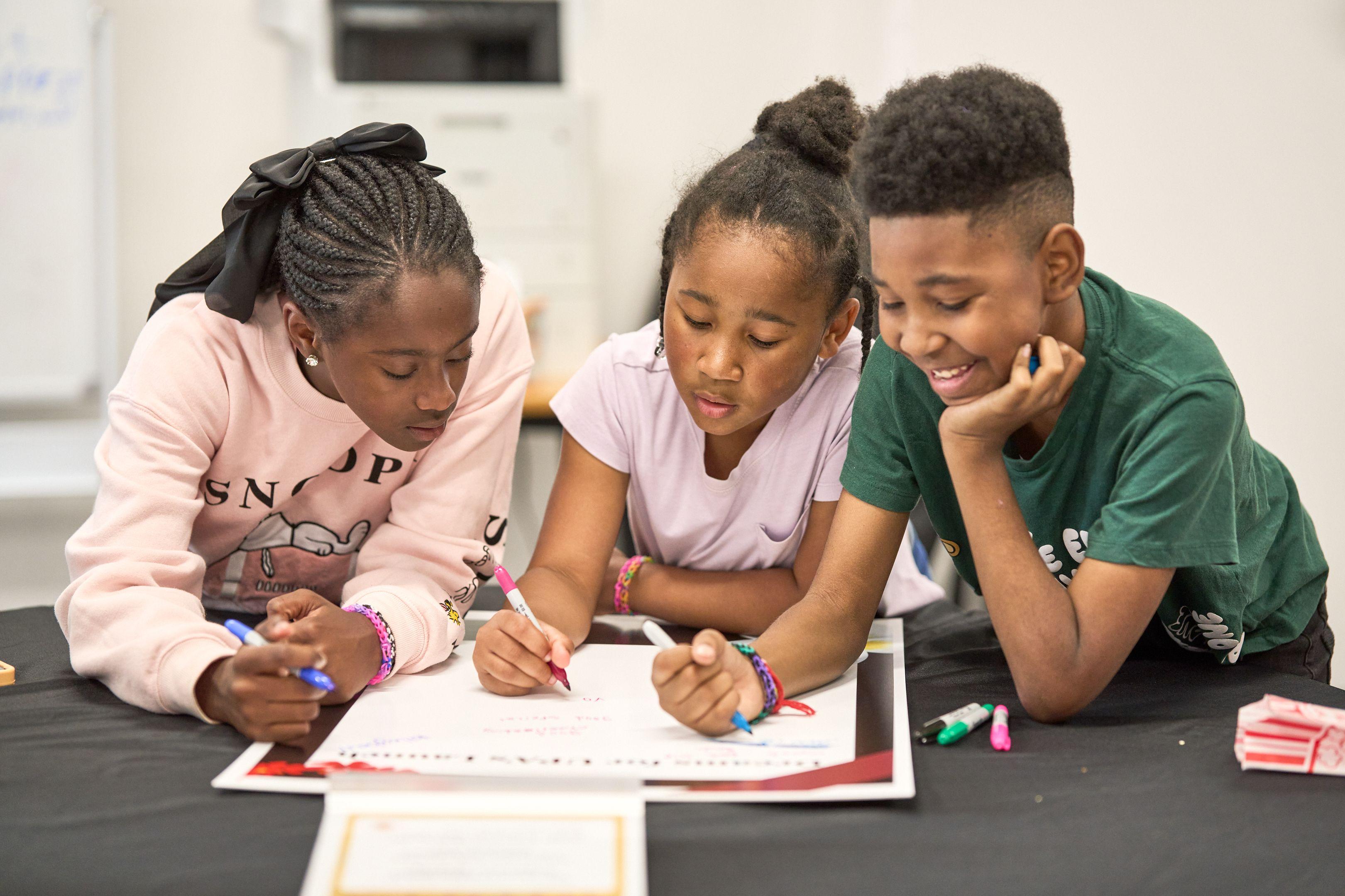 Three students leaning over a workbook together at a desk, writing and talking — School event photography, Austin Three students leaning over a workbook together at a desk, writing and talking