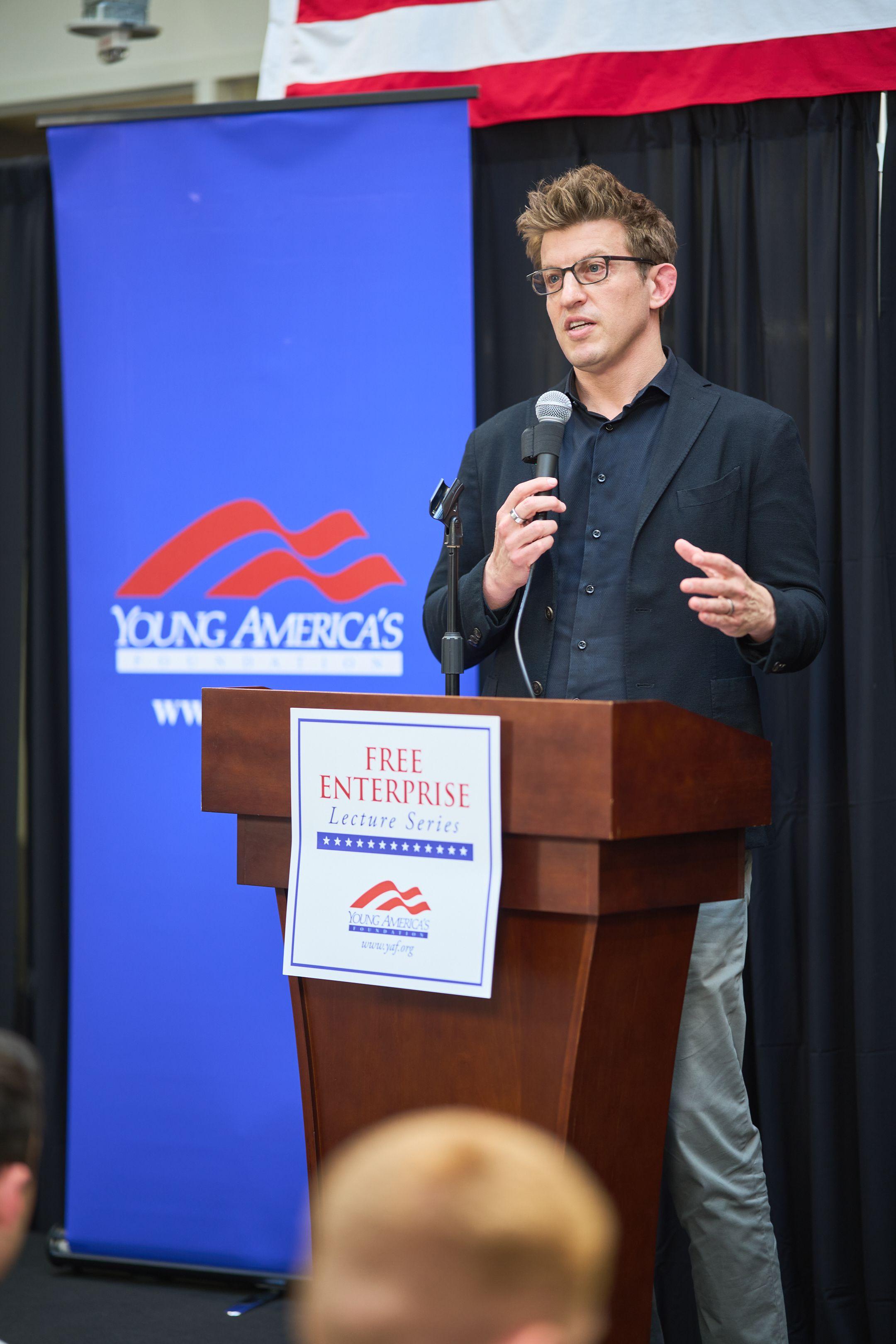 Speaker in glasses at a lectern with a microphone, addressing an audience — Production behind-the-scenes, Austin Speaker in glasses at a lectern with a microphone, addressing an audience in front of a blue backdrop