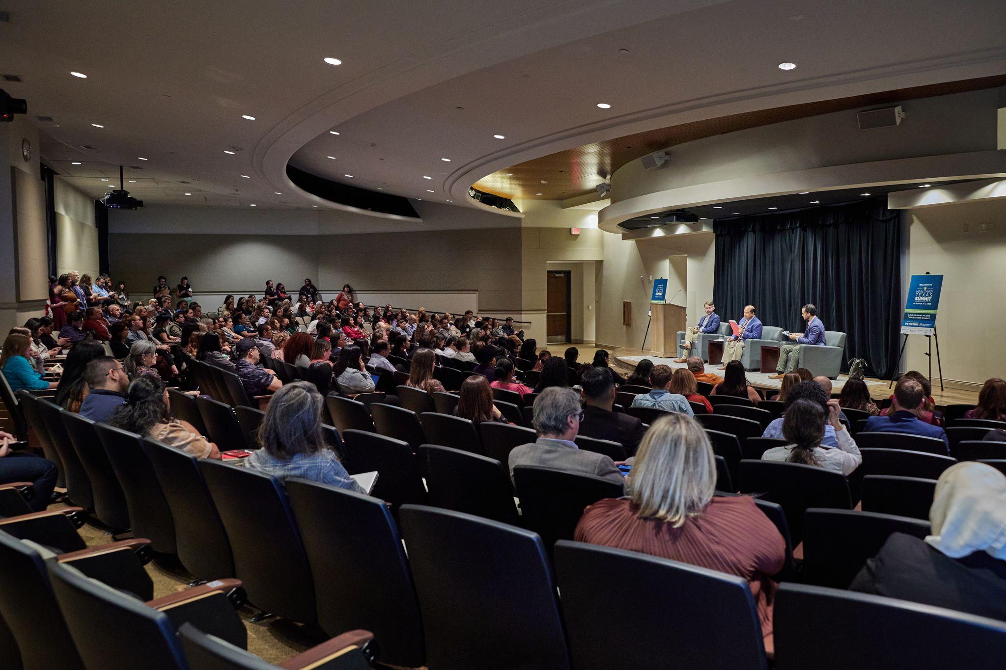 Full auditorium of seated delegates facing a lit conference stage — Conventions & Tradeshows photography, Austin A full auditorium of seated delegates facing a lit conference stage.