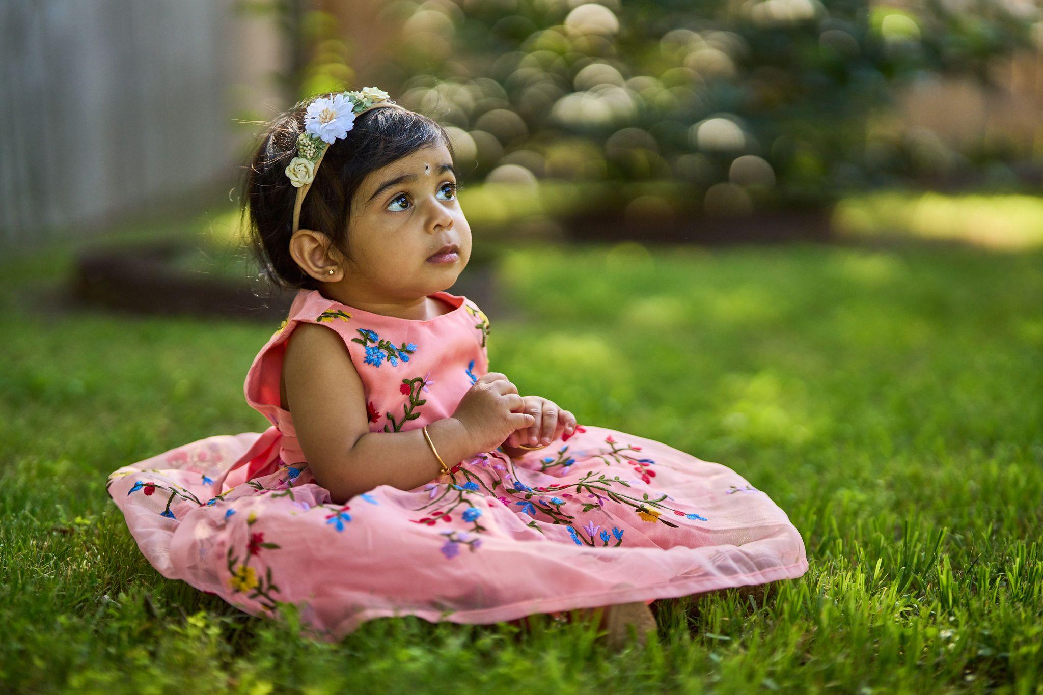 Toddler in a pink floral dress sitting on the grass, looking up — Kids Portraits photography, Austin A toddler in a pink floral dress sitting on the grass, looking up.