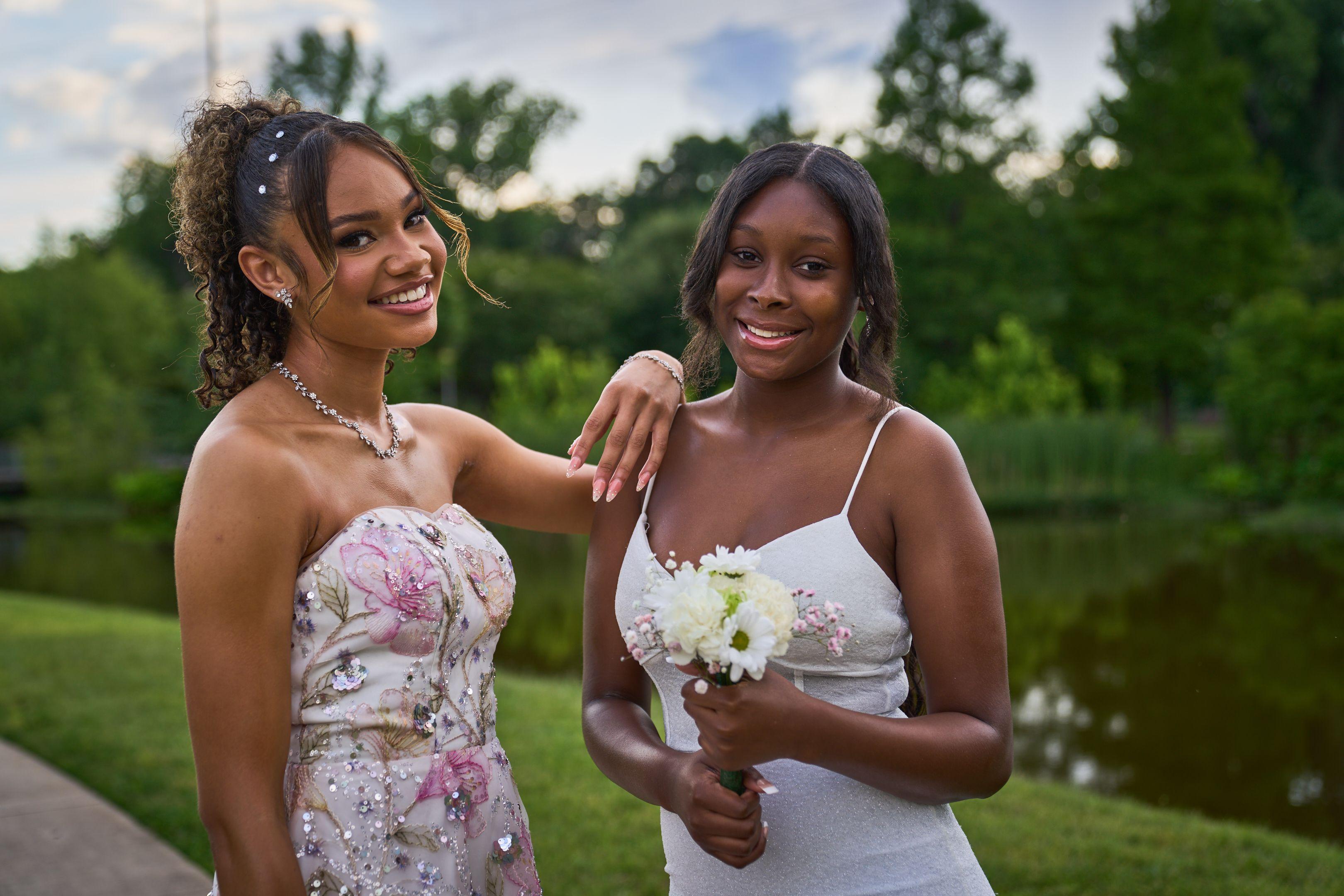 Two friends in prom dresses standing together on a lawn, one holding white flowers — Butler Metro Park photography, Austin Two friends in prom dresses standing together on a lawn, one holding white flowers.