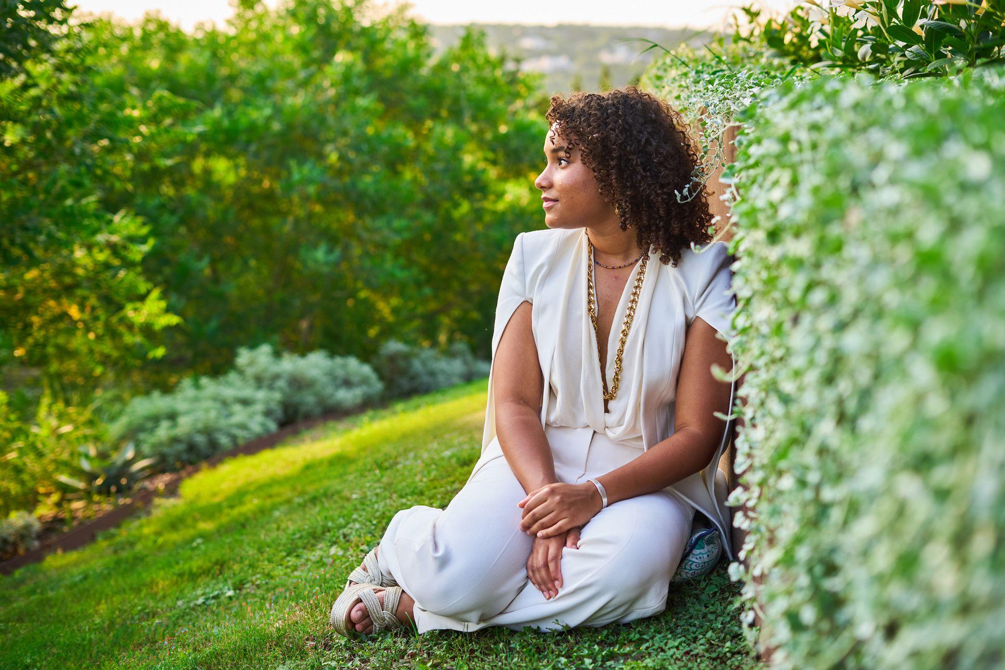 Woman in a white outfit seated on a lawn with greenery — Portrait photography, Austin A woman in a white outfit seated on a lawn with greenery behind her.