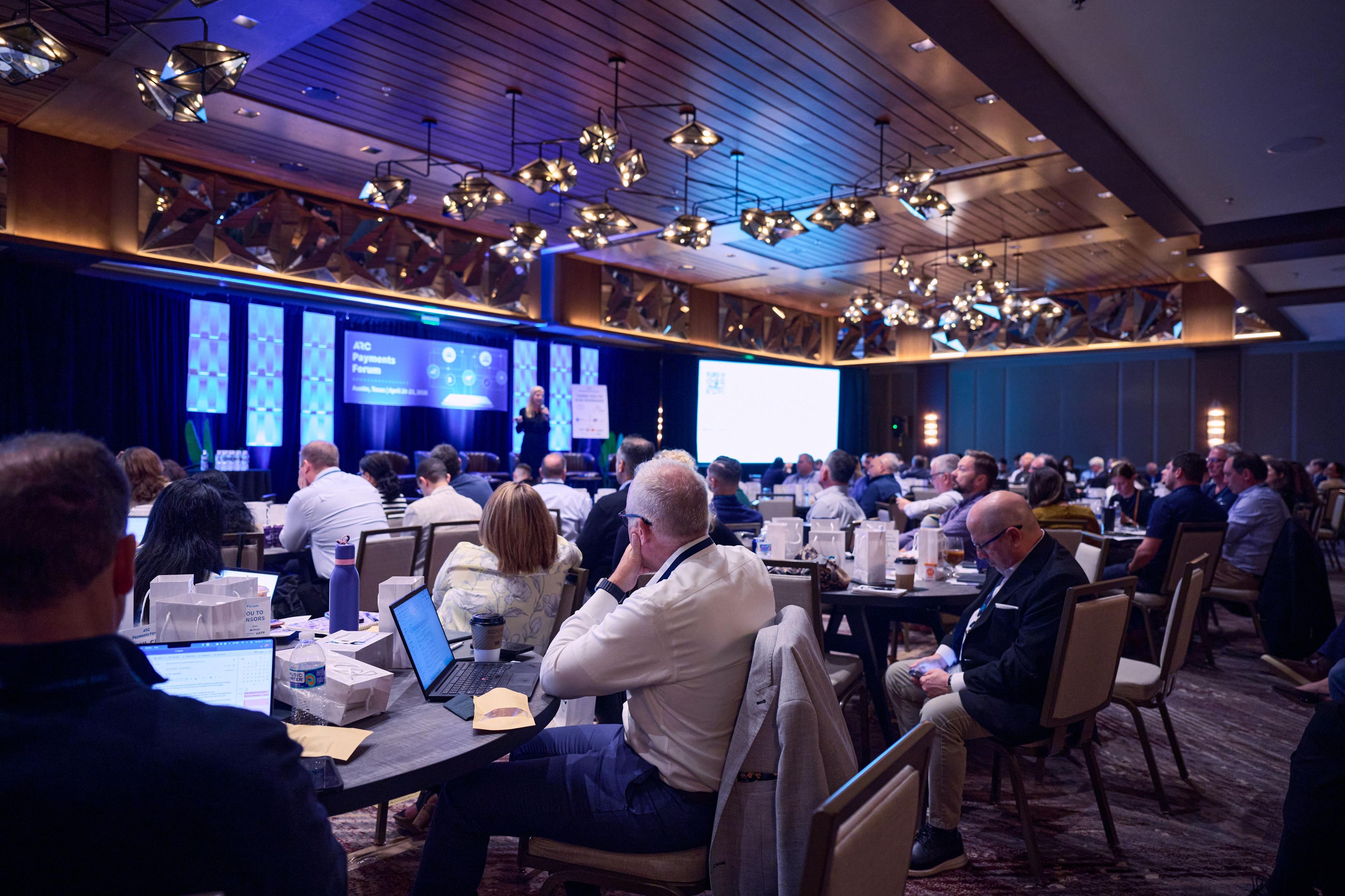 Packed conference ballroom at round tables, blue-lit screens across the front wall — Conventions & Tradeshows photography, Austin A packed conference ballroom at round tables, blue-lit screens across the front wall.