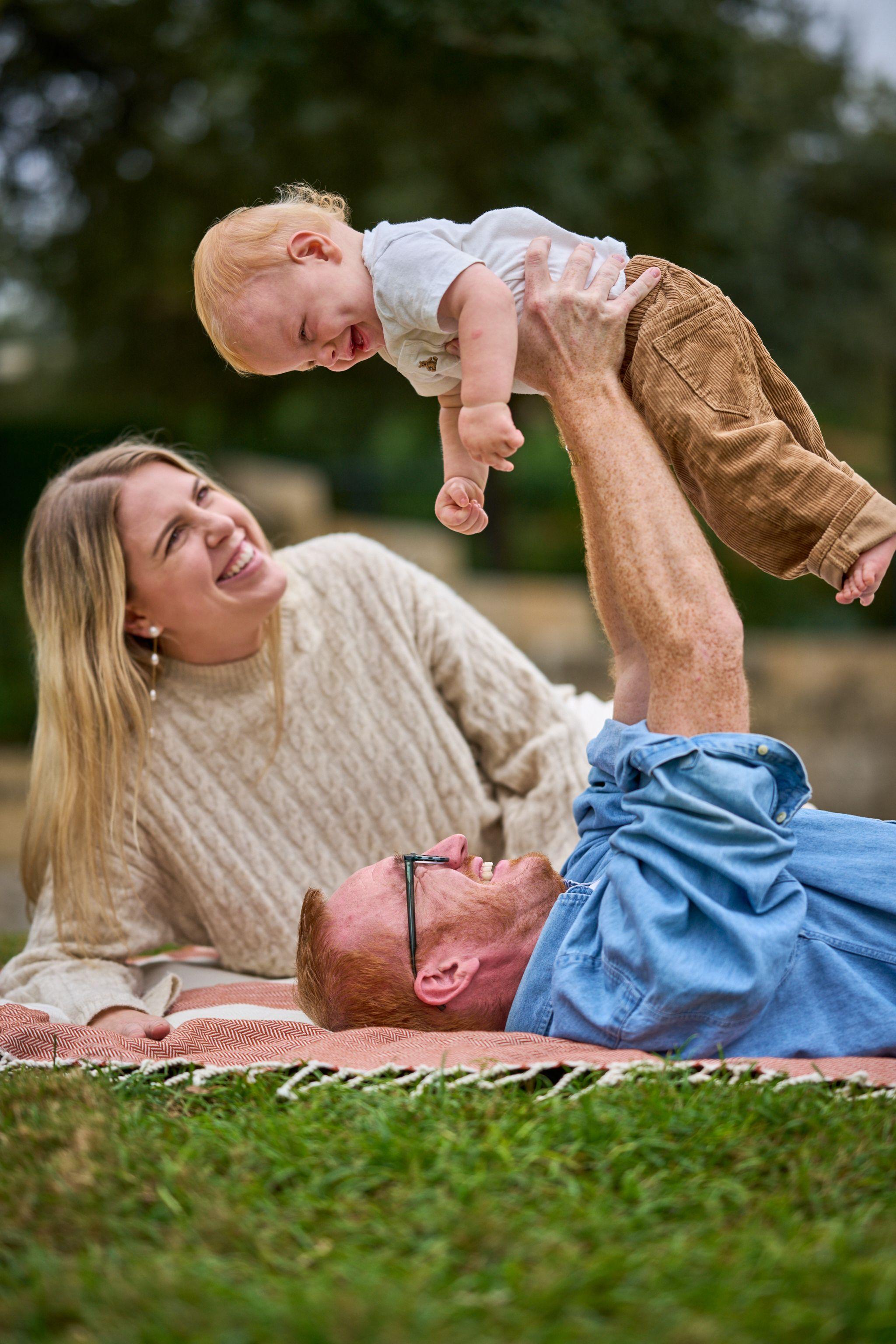 Father lying on a picnic blanket lifting his baby overhead while the mother laughs — Waterloo Park photography, Austin A father lying on a picnic blanket lifting his baby overhead while the mother laughs.