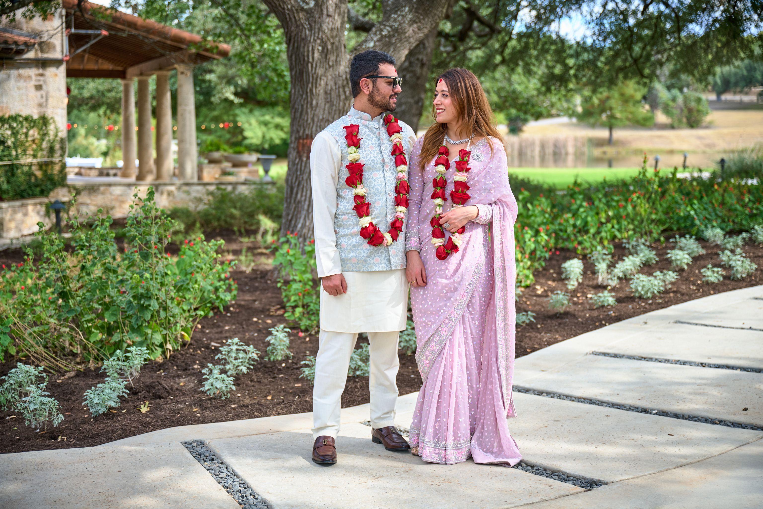 Couple in Indian wedding dress — a pink embroidered sari and white kurta — Weddings photography, Austin A couple in Indian wedding dress — a pink embroidered sari and white kurta with red garlands — standing on a garden path.