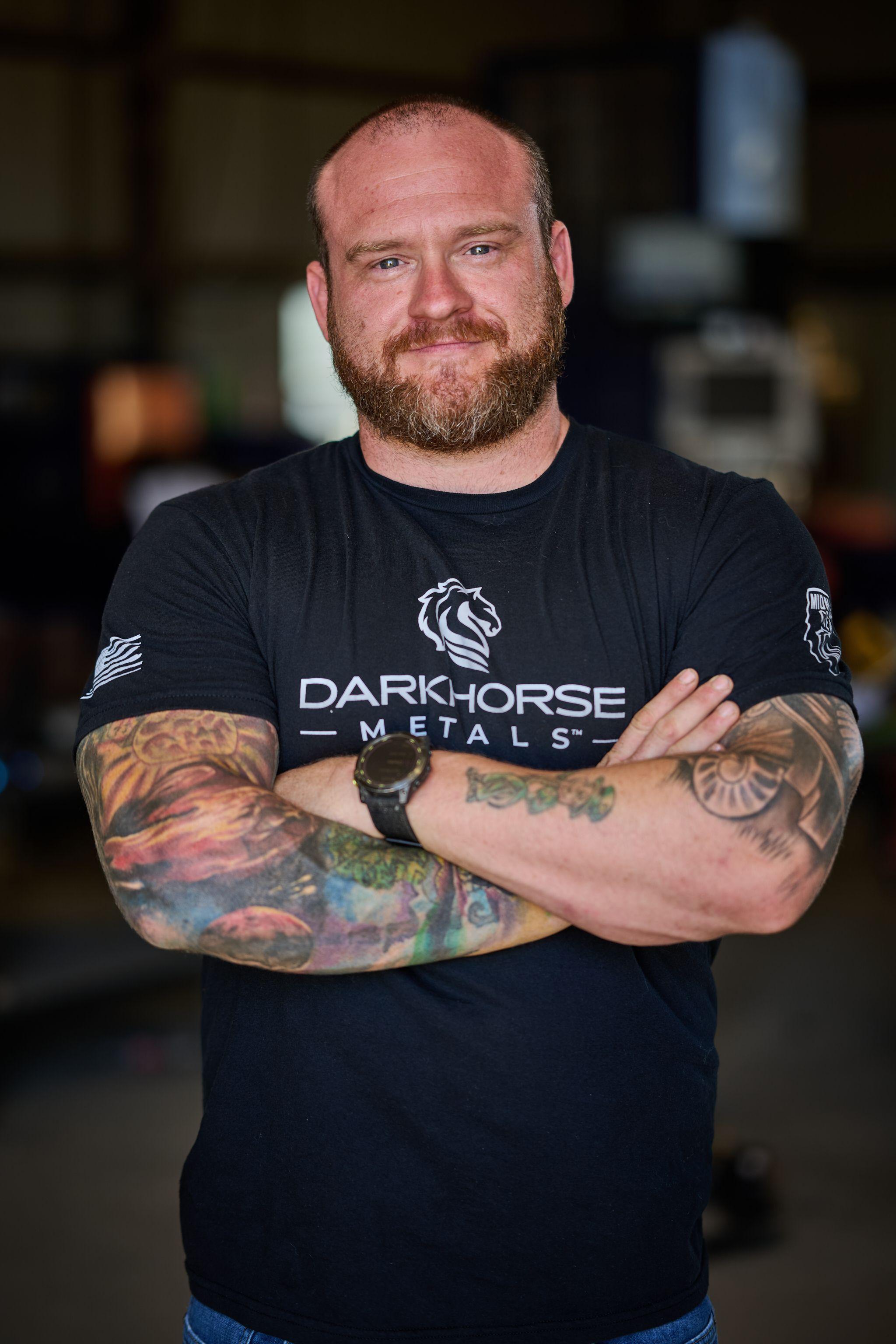 Tattooed man in a black metalworking company t-shirt standing with his arms crossed — Business headshots, Austin A tattooed man in a black metalworking company t-shirt standing with his arms crossed in a workshop