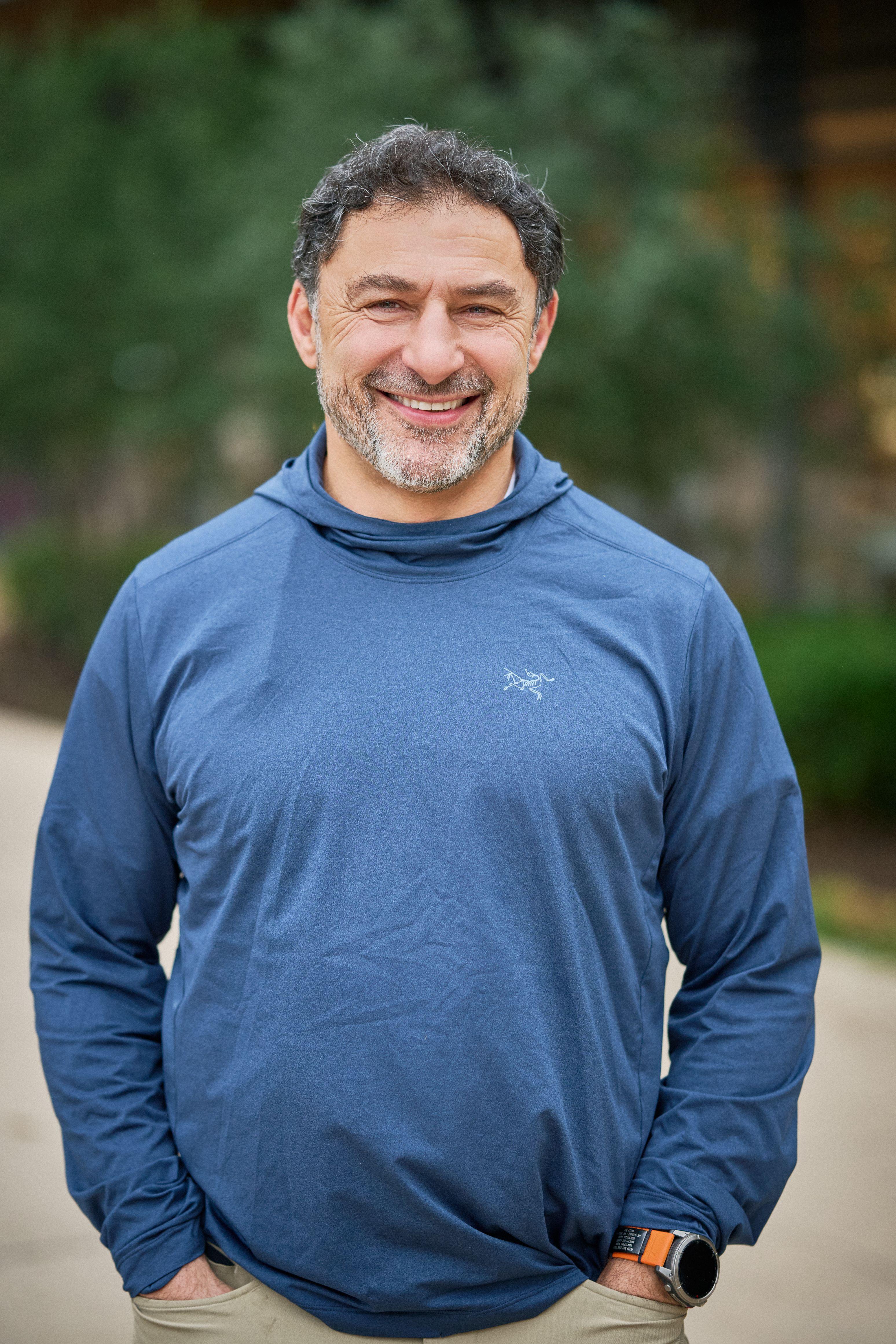 Older man with gray hair and a beard in a blue technical top, photographed outdoors — Business headshots, Austin An older man with gray hair and a beard in a blue technical top, photographed outdoors with greenery behind him