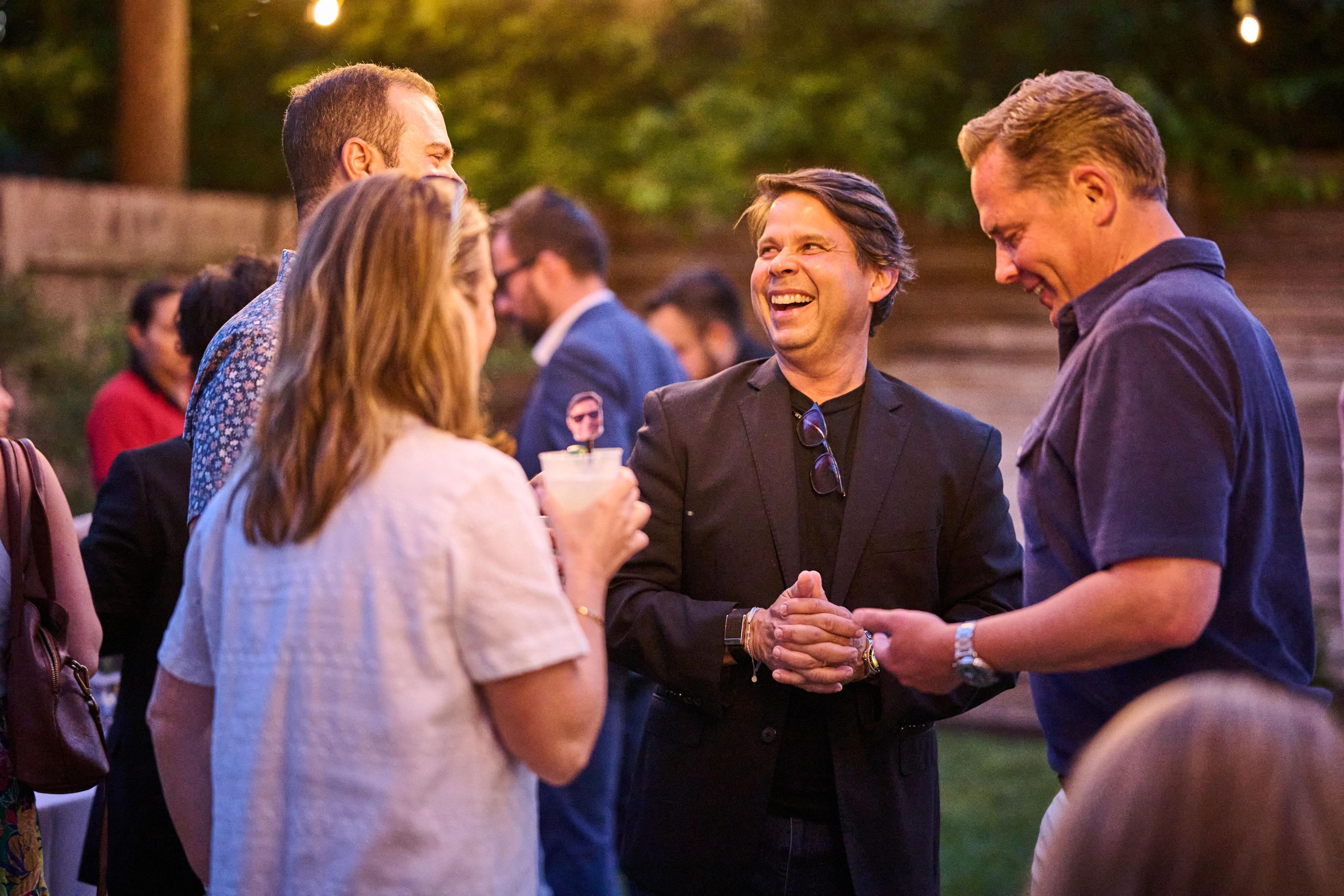 Man in a dark suit laughing among guests at an outdoor evening reception — Formal Celebrations photography, Austin A man in a dark suit laughing among guests at an outdoor evening reception.