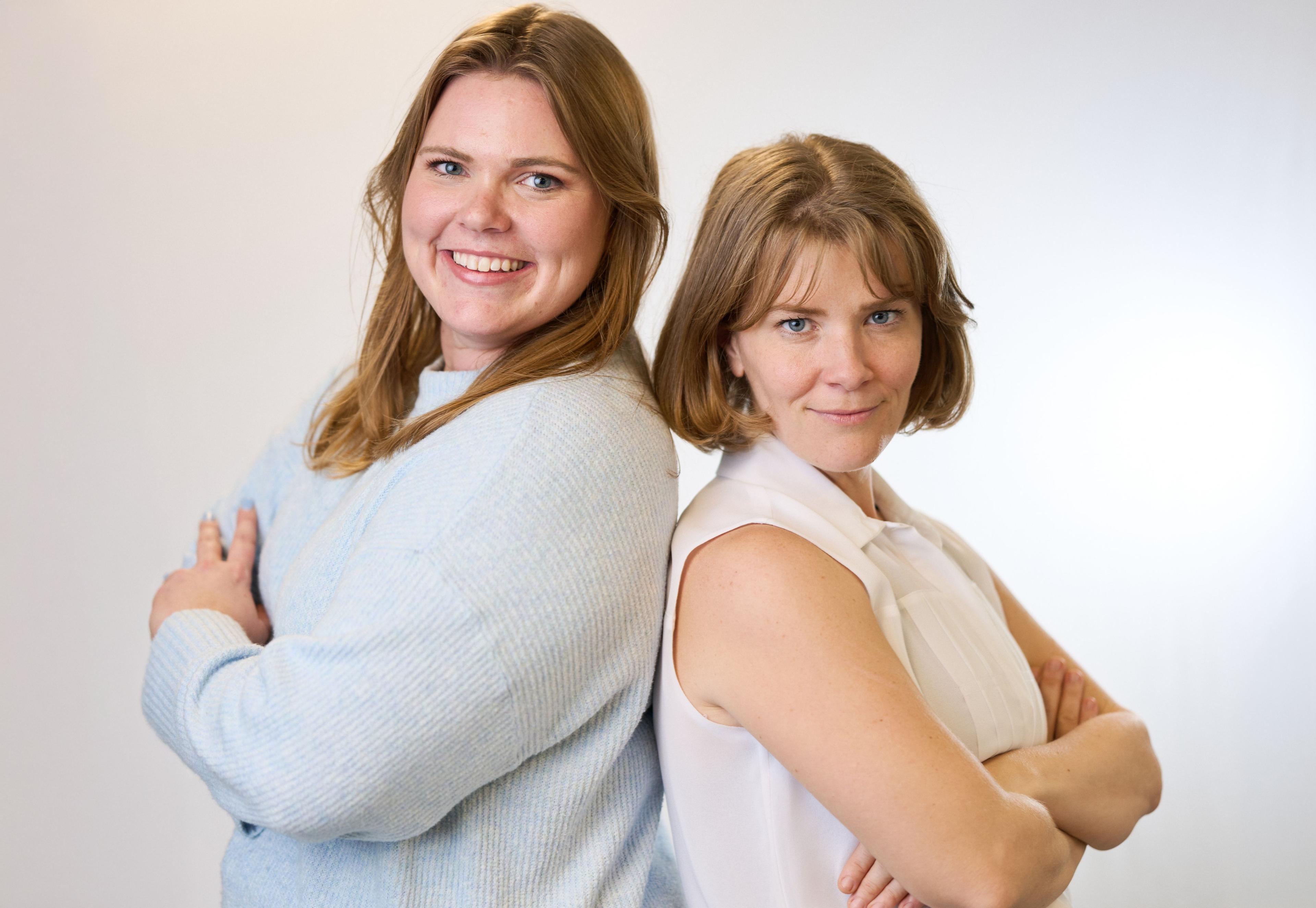 Two women standing back to back, one smiling at the camera, on a light studio background — Commercial photo studio, Austin Two women standing back to back, one smiling at the camera, on a light studio background