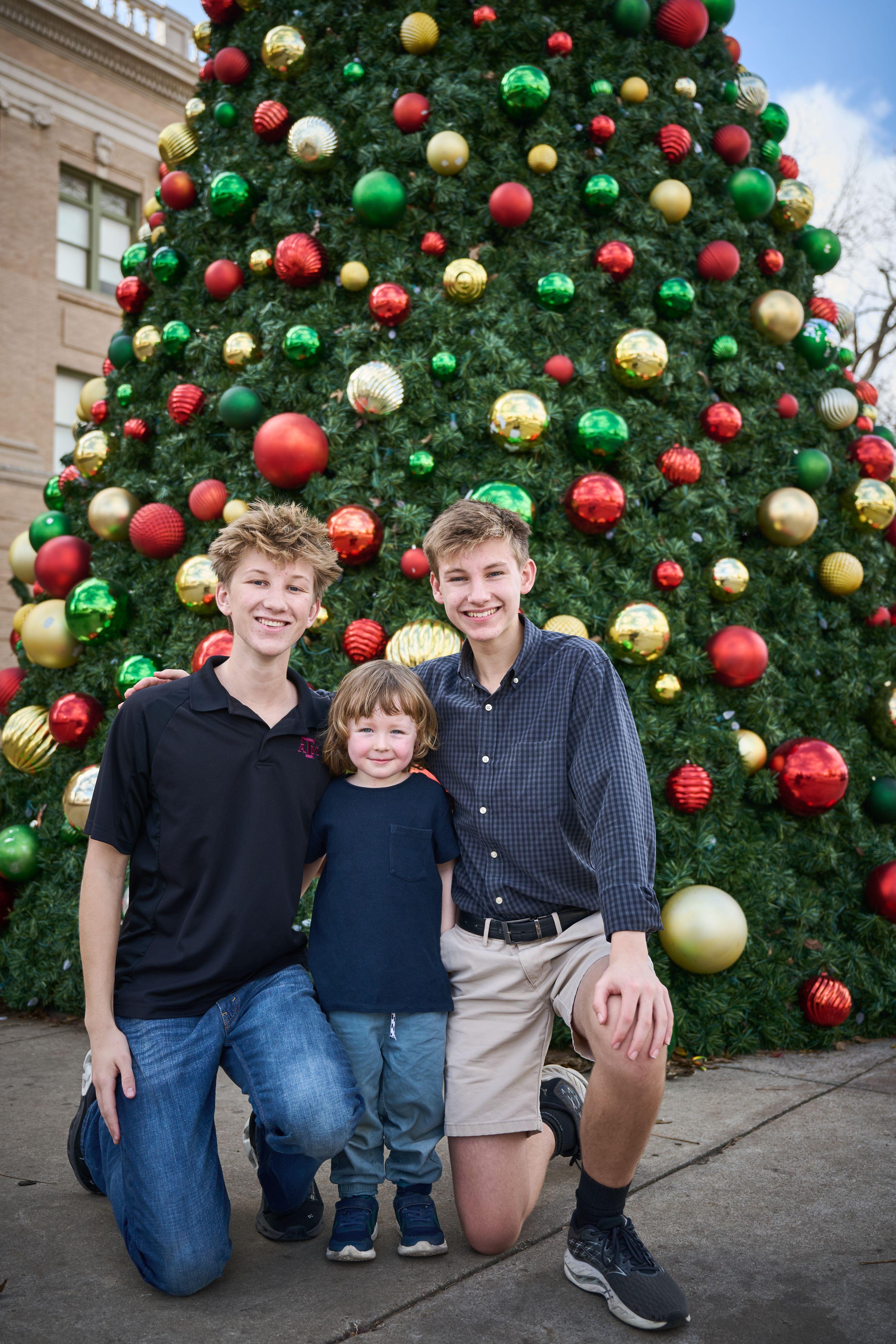 Three brothers and their father crouched together at the foot of a giant Christmas tree — Georgetown Downtown photography, Austin Three brothers and their father crouched together at the foot of a giant Christmas tree.