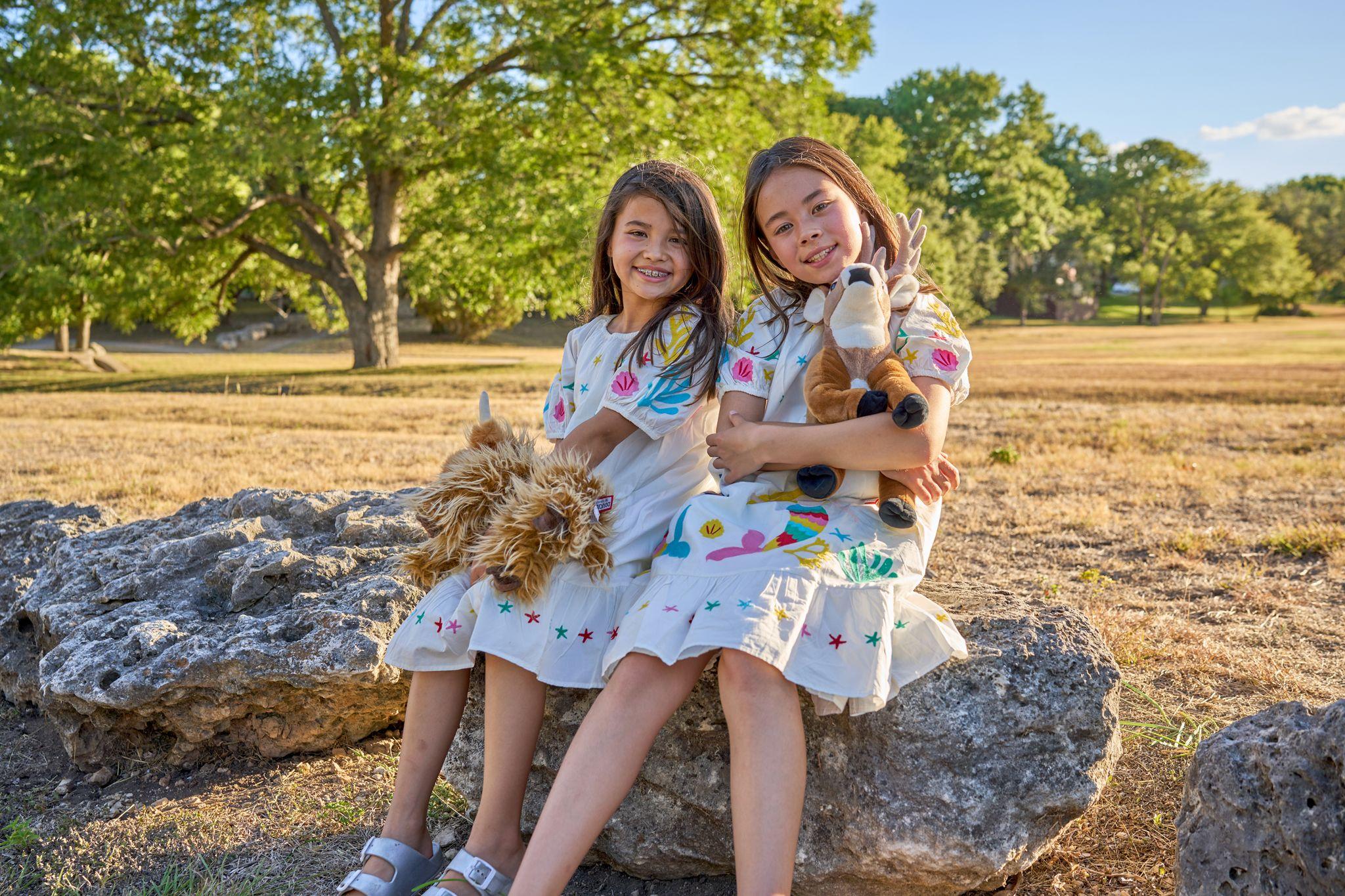 Two sisters in embroidered dresses sitting on a rock holding stuffed toys — Kids Portraits photography, Austin Two sisters in embroidered dresses sitting on a rock holding stuffed toys.