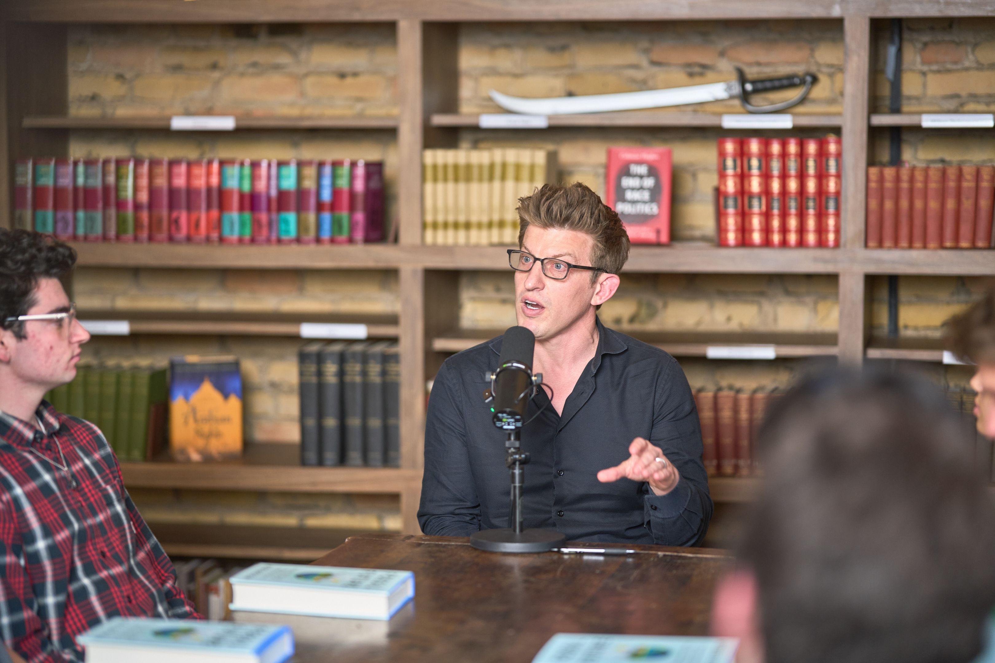 Speaker in glasses talking at a wooden table in a book-lined library room — Production behind-the-scenes, Austin Speaker in glasses talking at a wooden table in a book-lined library room