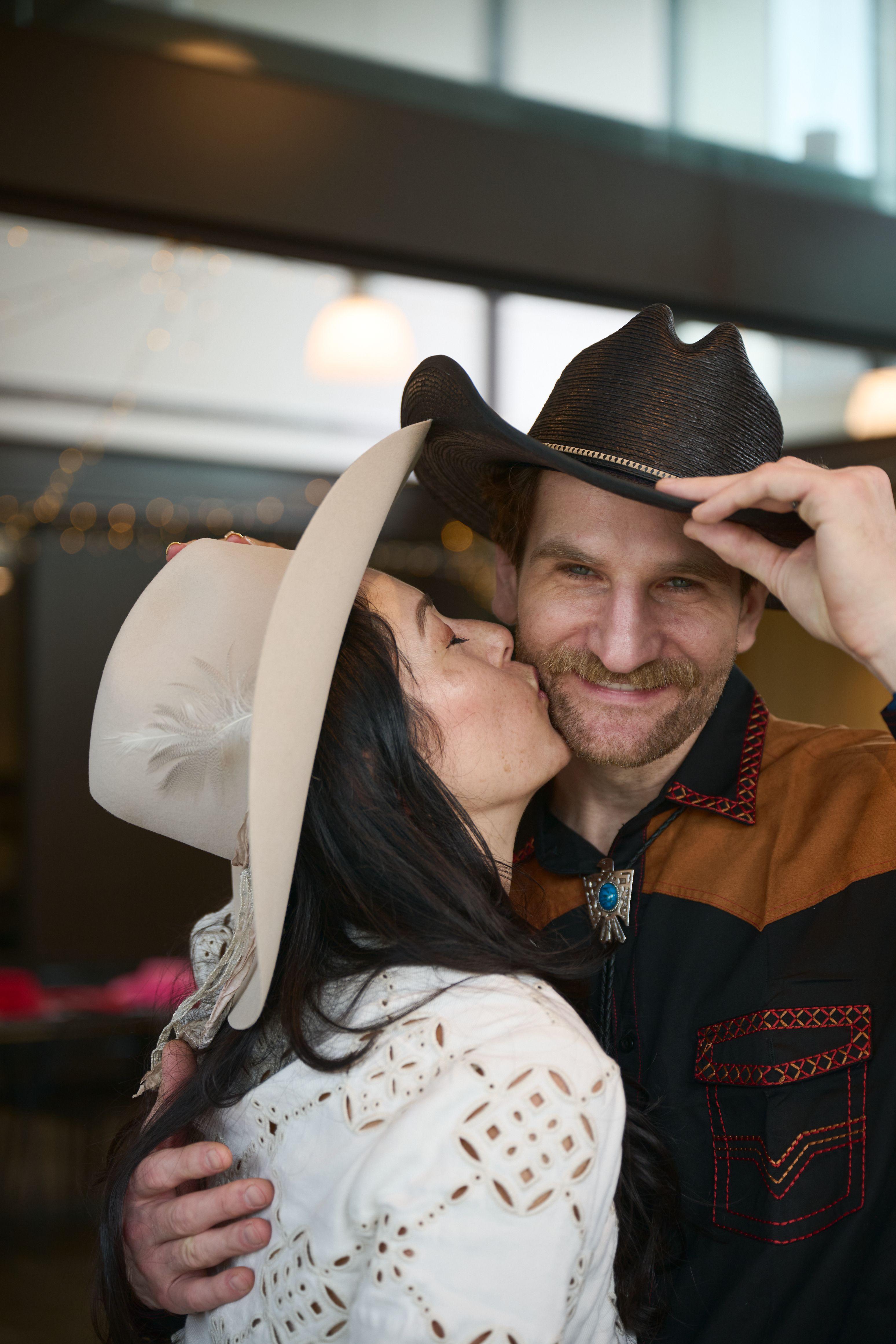Woman kissing her partner's cheek as he tips a cowboy hat, both in western shirts — Couples & Anniversary photography, Austin A woman kissing her partner's cheek as he tips a cowboy hat, both in western shirts.