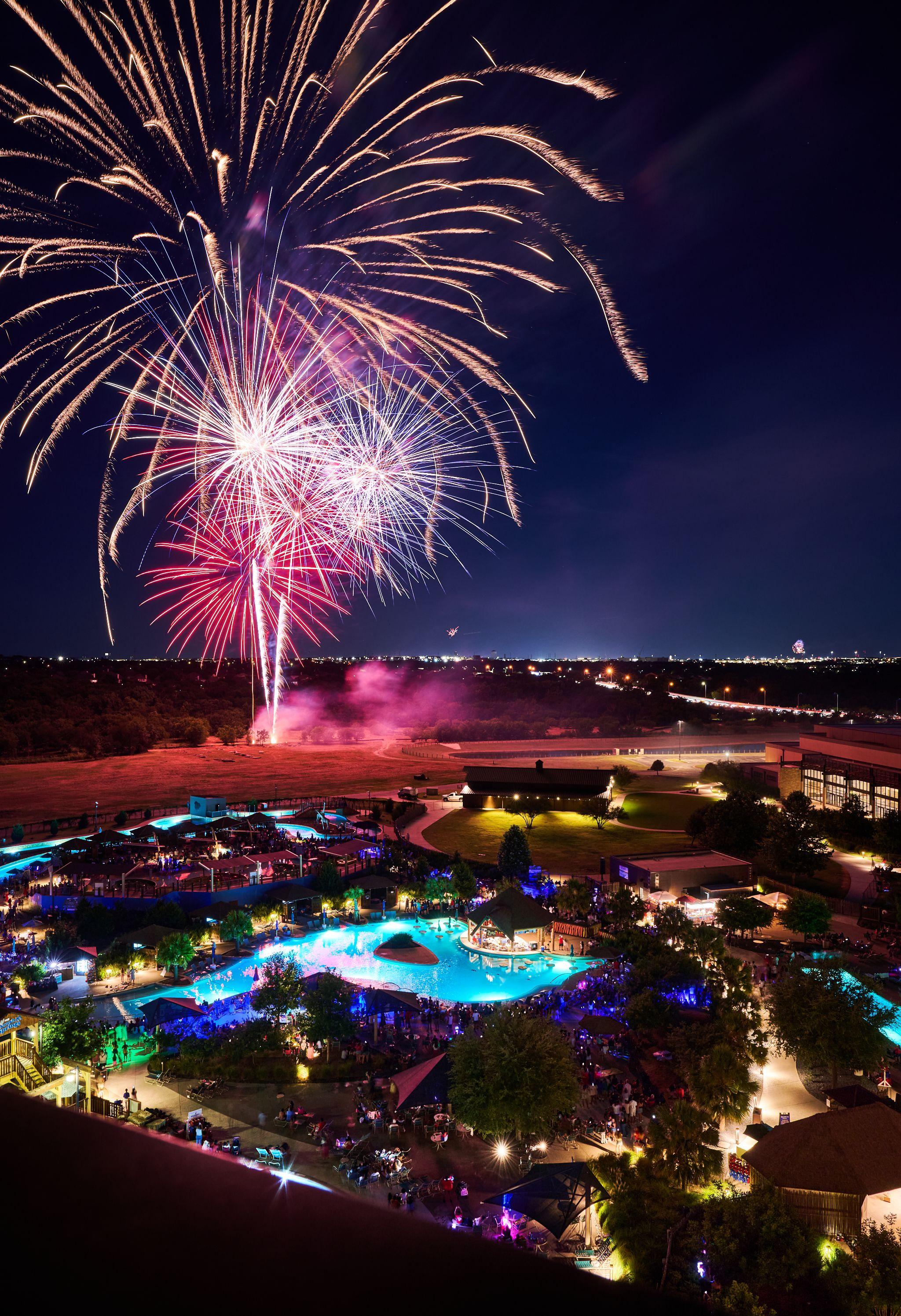 Fireworks bursting pink and white above a lit waterpark at night — Festival photography, Austin Fireworks bursting pink and white above a lit waterpark at night.