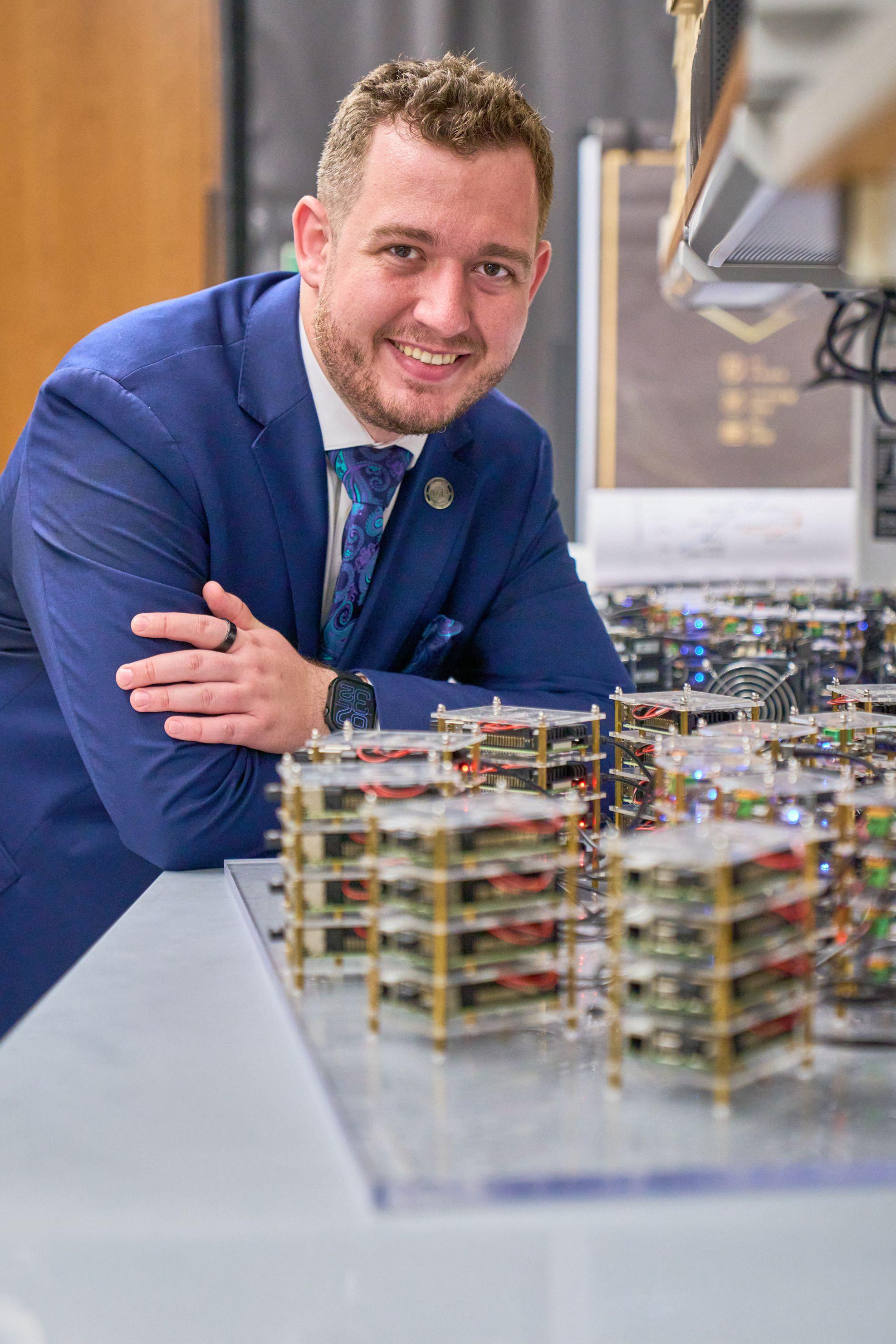 Man in a blue suit leaning on a bench beside stacked electronics in a campus lab — UT Austin photography, Austin A man in a blue suit leaning on a bench beside stacked electronics in a campus lab.