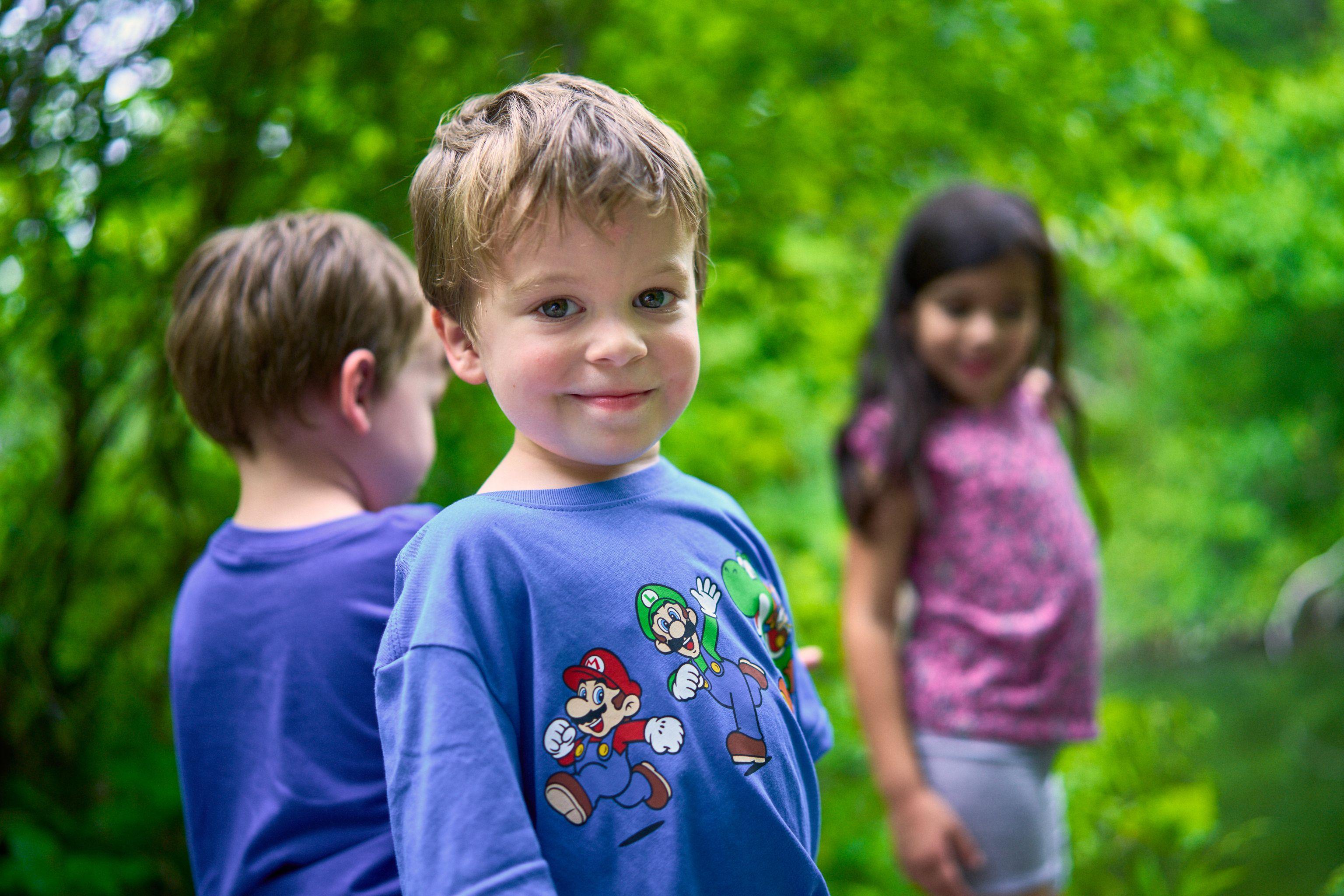Young boy in a Mario T-shirt smiling at camera, siblings blurred either side — Family Portraits photography, Austin A young boy in a Mario T-shirt smiling at camera, siblings blurred either side.