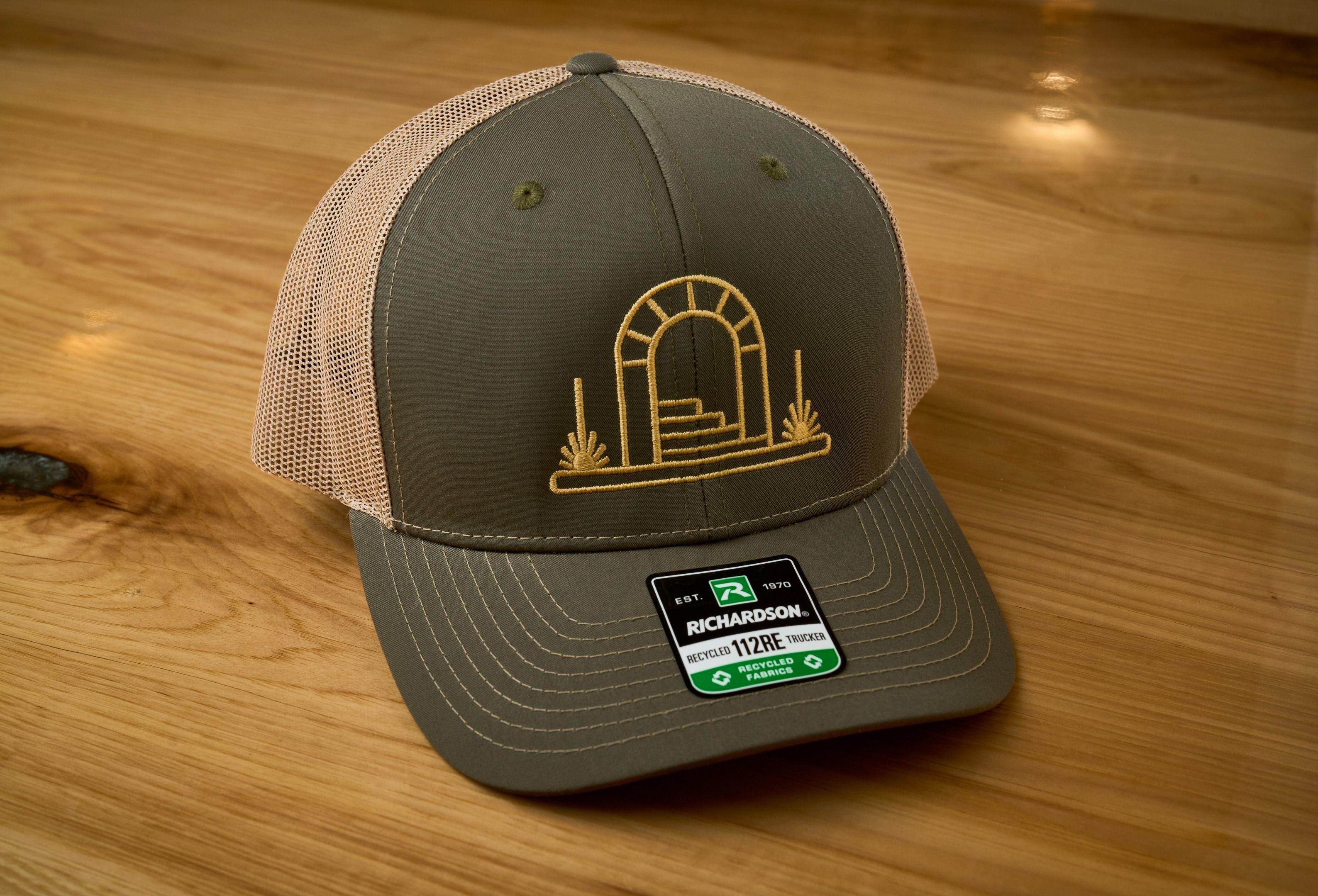 Olive trucker cap with an embroidered arch logo on a wooden surface — Product photography, Austin An olive trucker cap with an embroidered arch logo on a wooden surface.