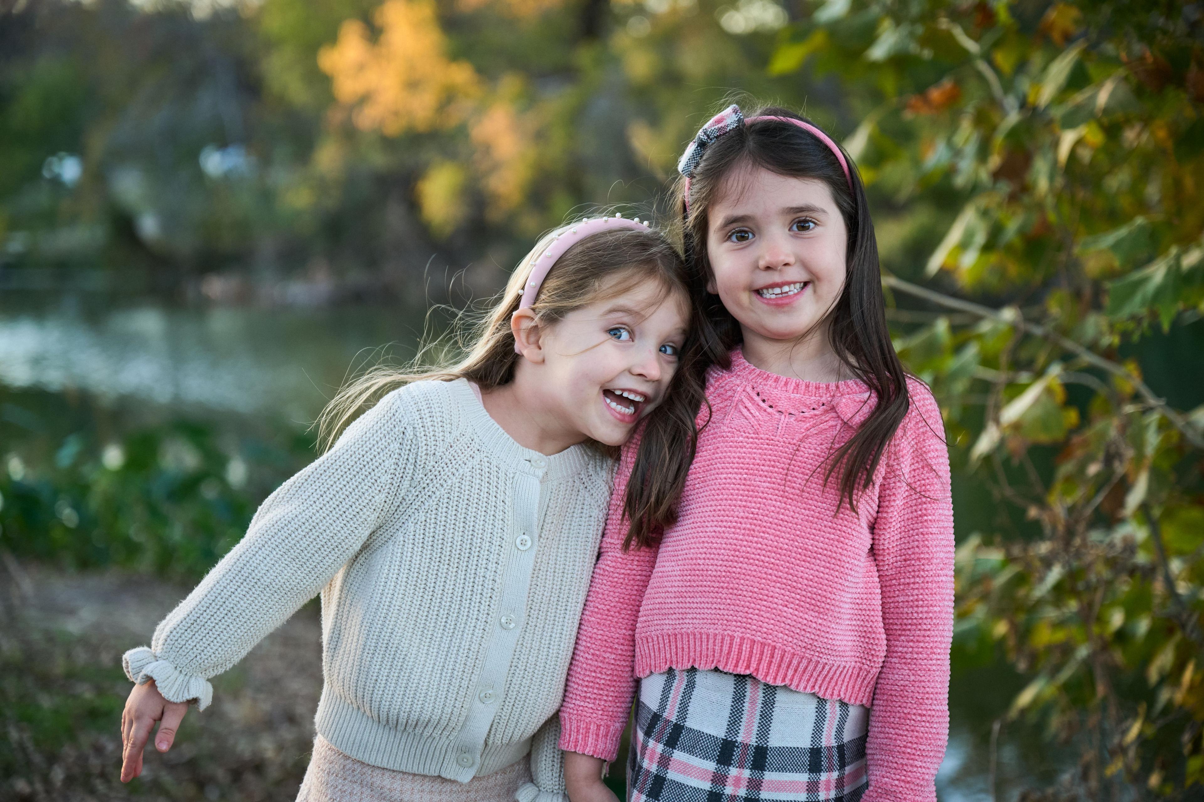 Two sisters in pink and cream knitwear leaning together, autumn trees — Family Portraits photography, Austin Two sisters in pink and cream knitwear leaning together, autumn trees behind.