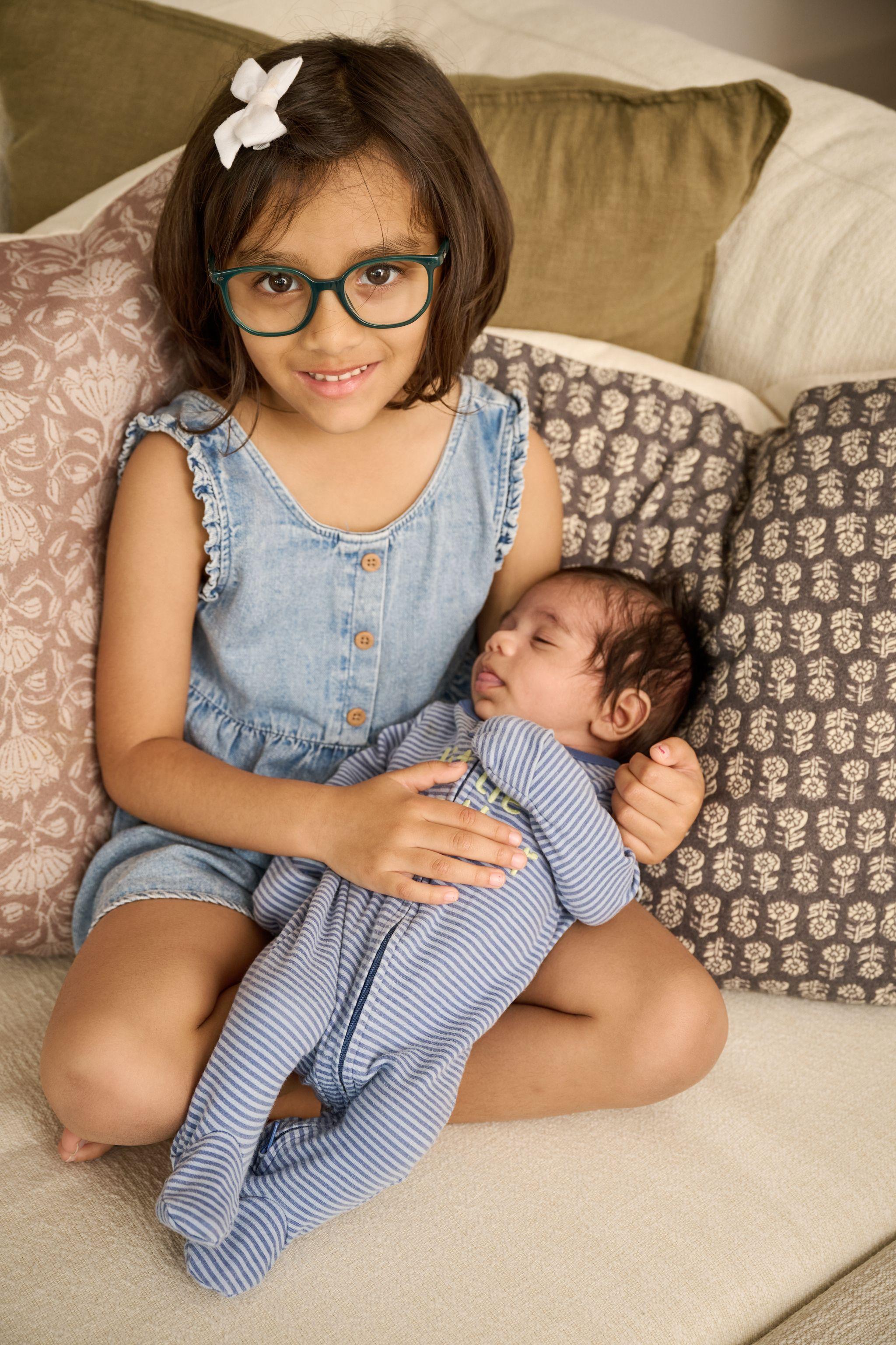 Young girl in a denim dress sitting beside her newborn sibling on a sofa — Newborn photography, Austin A young girl in a denim dress sitting beside her newborn sibling on a sofa.