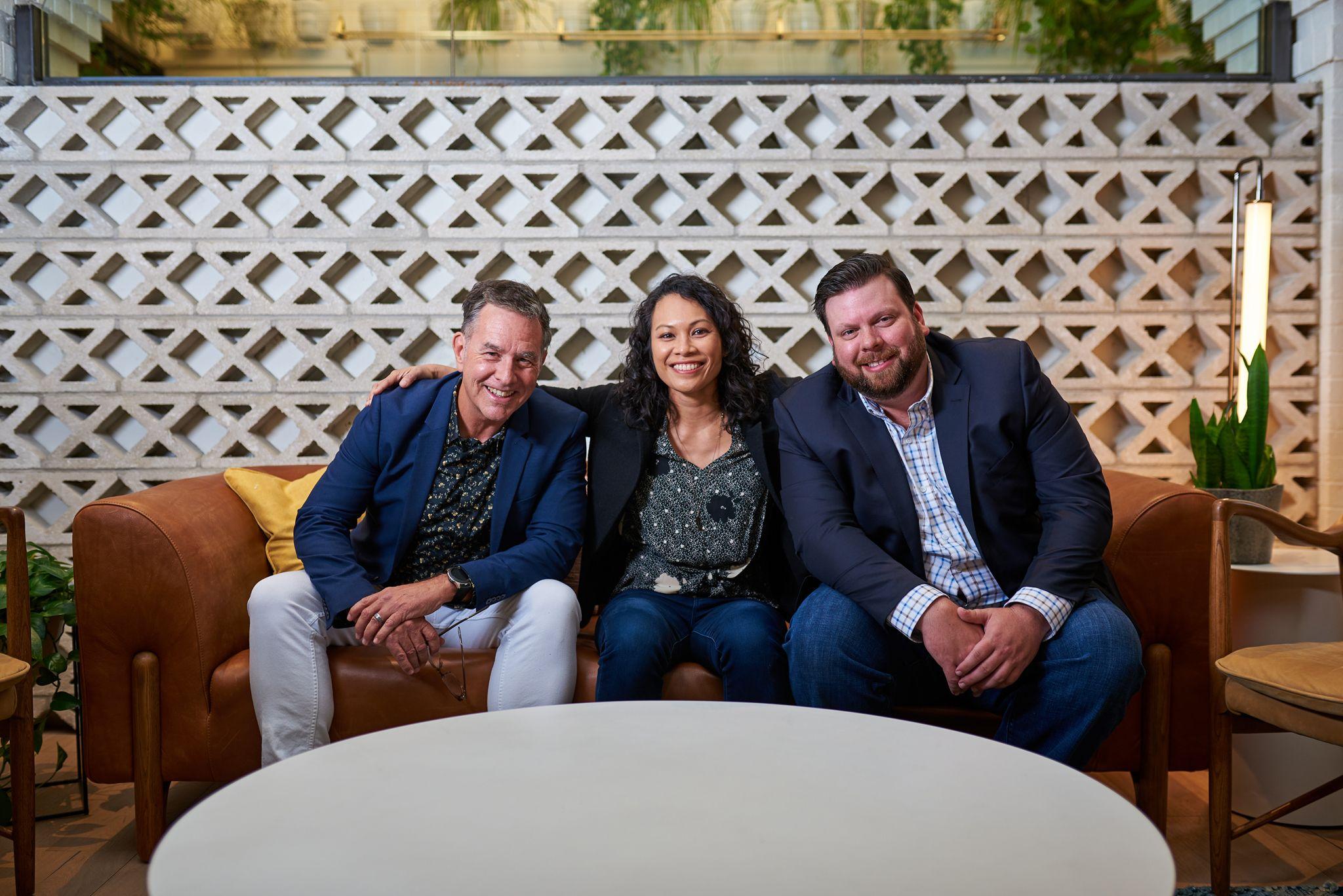 Three colleagues sitting together on a sofa in front of a white breeze-block wall — Team photography, Austin Three colleagues sitting together on a sofa in front of a white breeze-block wall