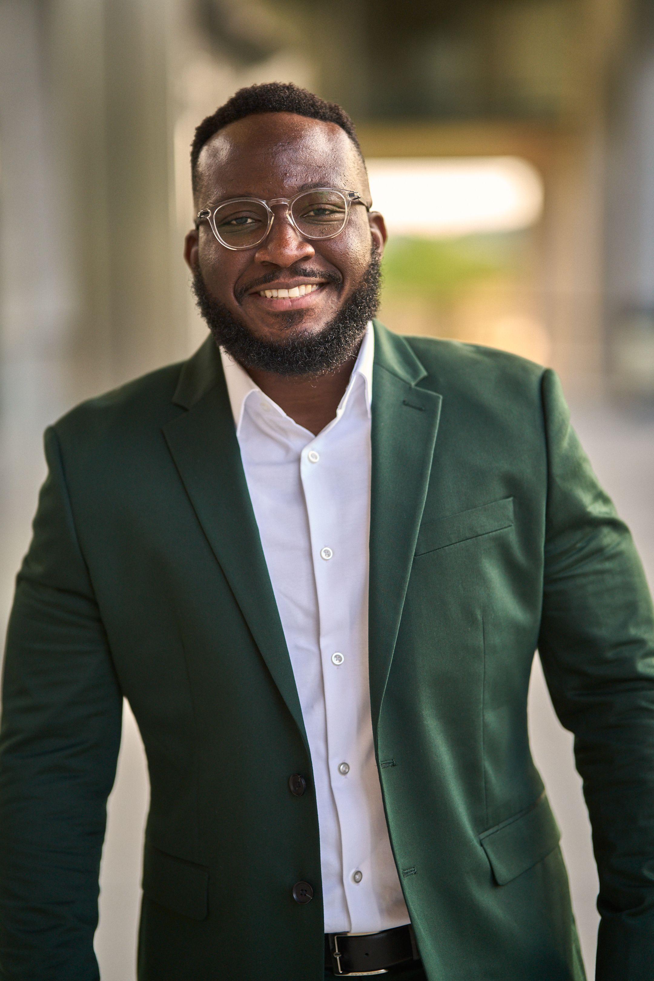 Man in a dark green blazer and open white shirt smiling in a business headshot, the building — Business headshots, Austin A man in a dark green blazer and open white shirt smiling in a business headshot, the building behind him thrown out of focus