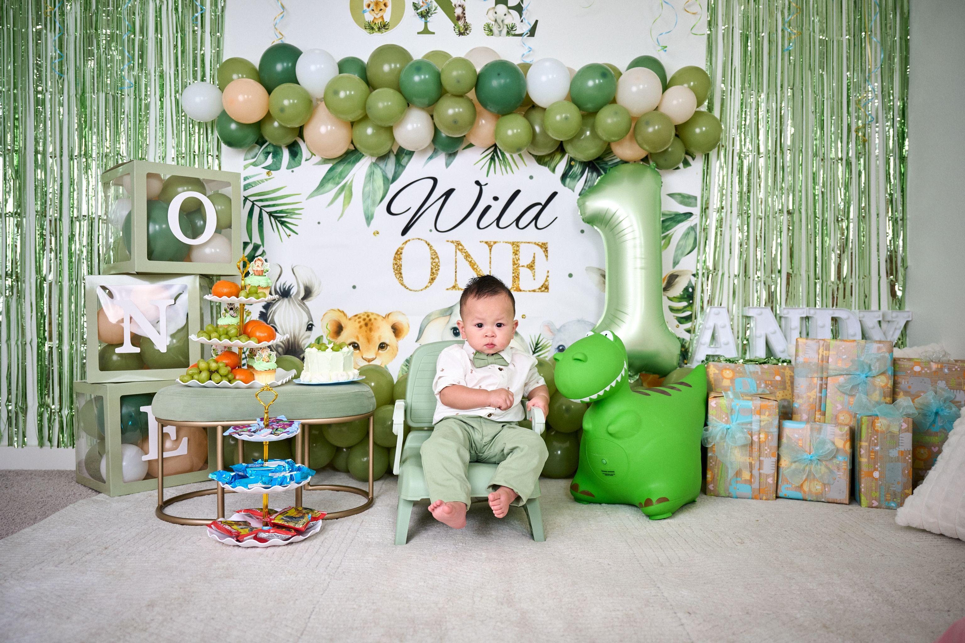 Baby in a green outfit sitting on a small chair — Family Parties photography, Austin A baby in a green outfit sitting on a small chair in front of a jungle-themed first birthday backdrop with a number one balloon