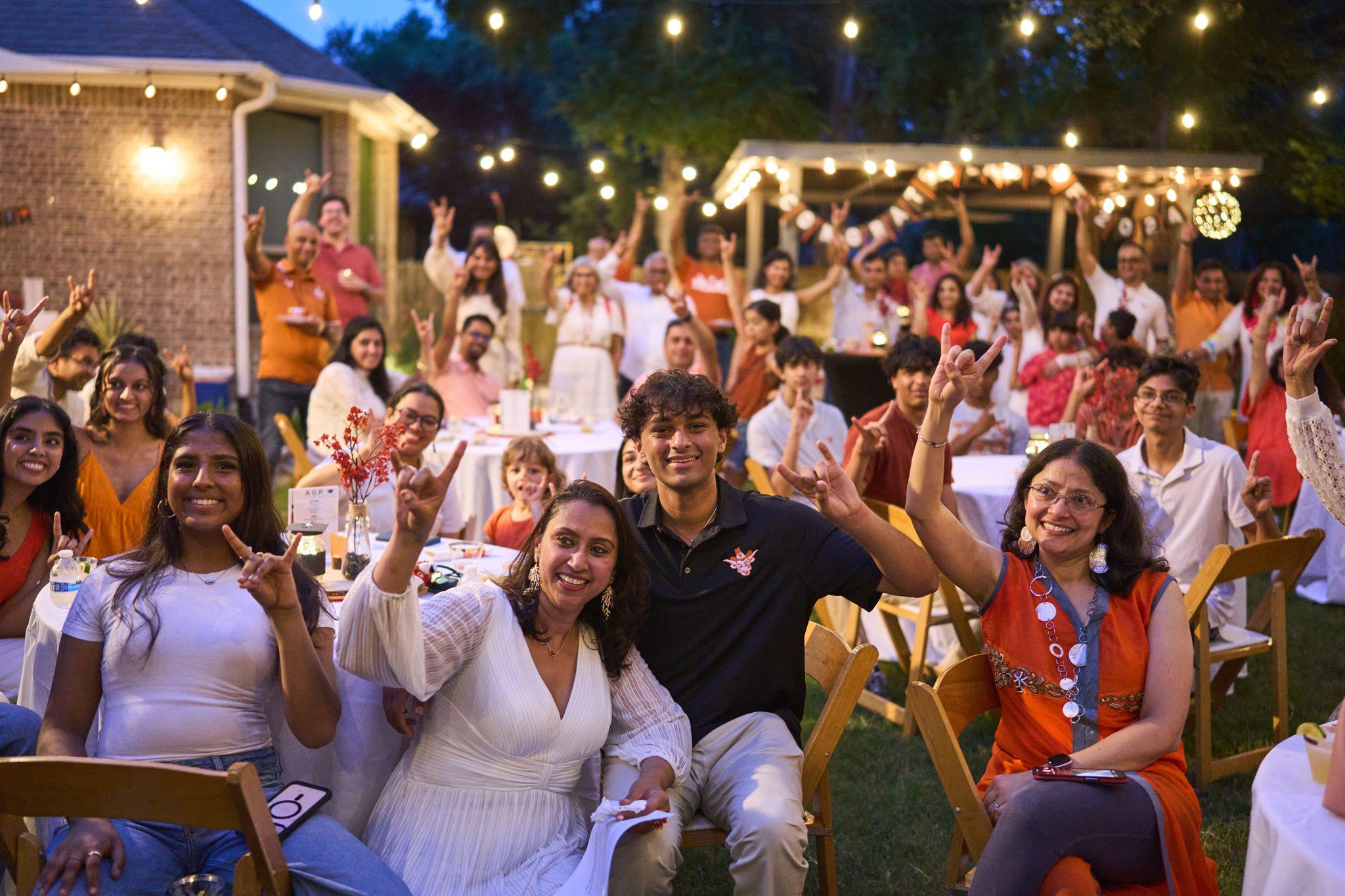 Guests at long tables under string lights raising their hands at a graduation party — Family Parties photography, Austin Guests at long tables under string lights raising their hands at a graduation party.