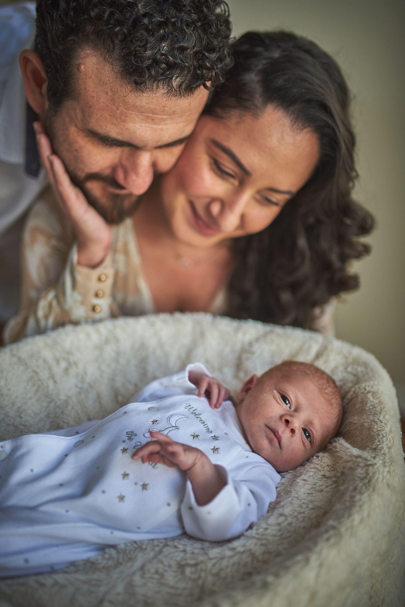 New parents leaning together over their newborn asleep in a soft basket — Newborn photography, Austin New parents leaning together over their newborn asleep in a soft basket.