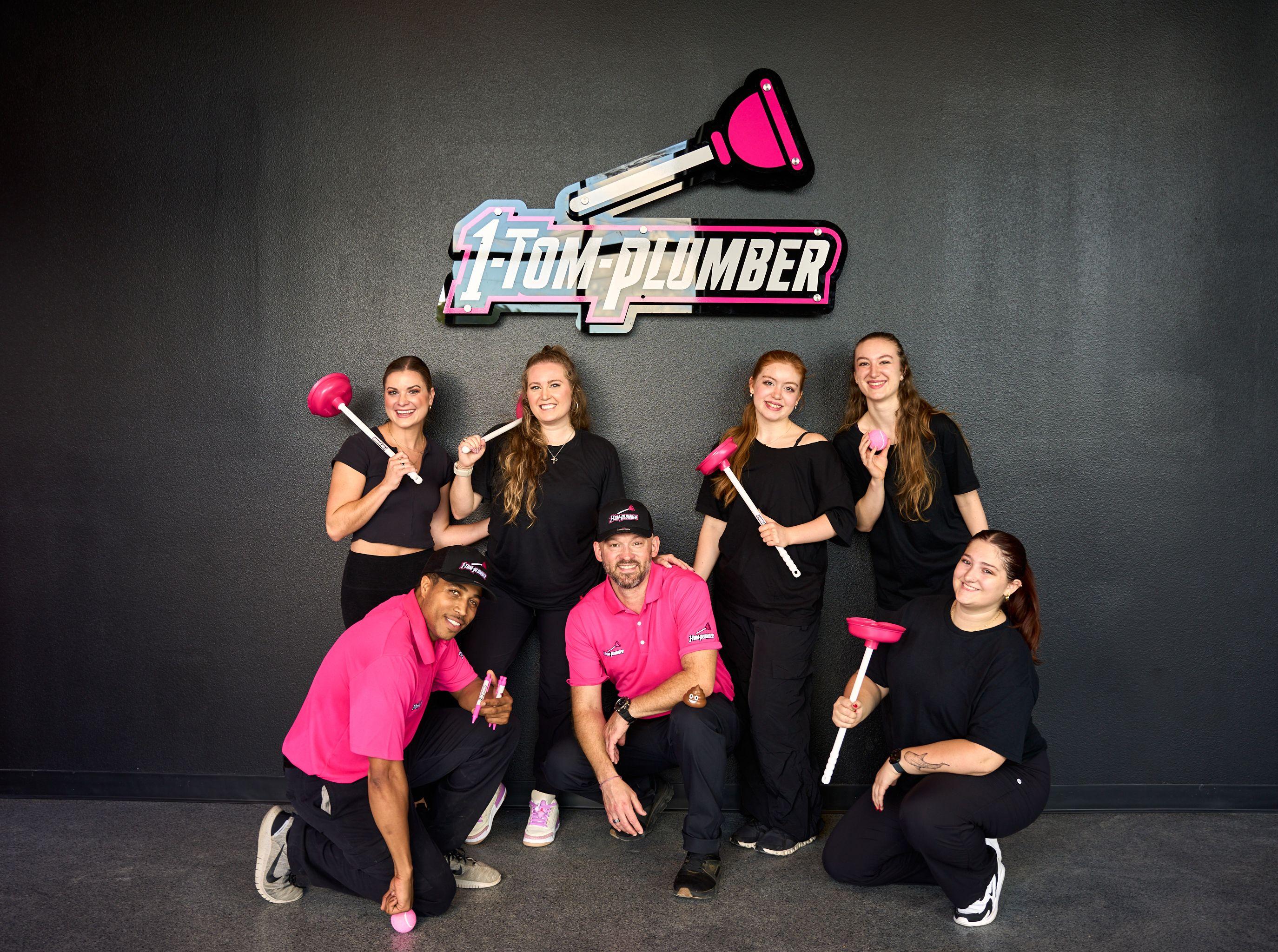 Team in black and pink posed playfully with plungers under a 1-Tom-Plumber sign — Team photography, Austin Team in black and pink posed playfully with plungers under a 1-Tom-Plumber sign