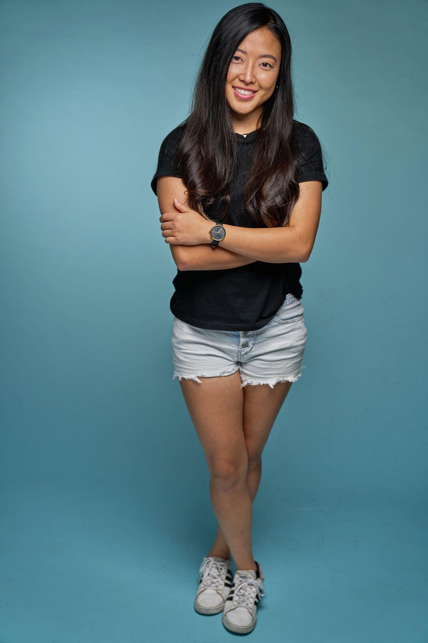 Young woman in a black tee and denim shorts standing full length against a teal studio backdrop — Commercial photo studio, Austin Young woman in a black tee and denim shorts standing full length against a teal studio backdrop