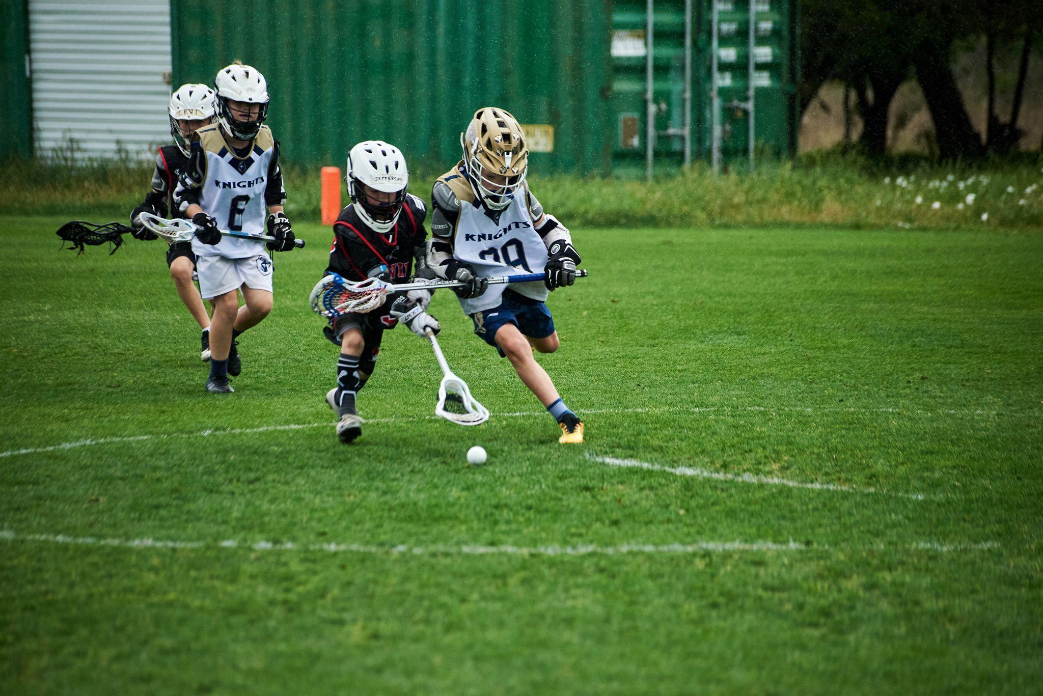 Youth lacrosse players in helmets and pads running for the ball on a grass field — School event photography, Austin Youth lacrosse players in helmets and pads running for the ball on a grass field