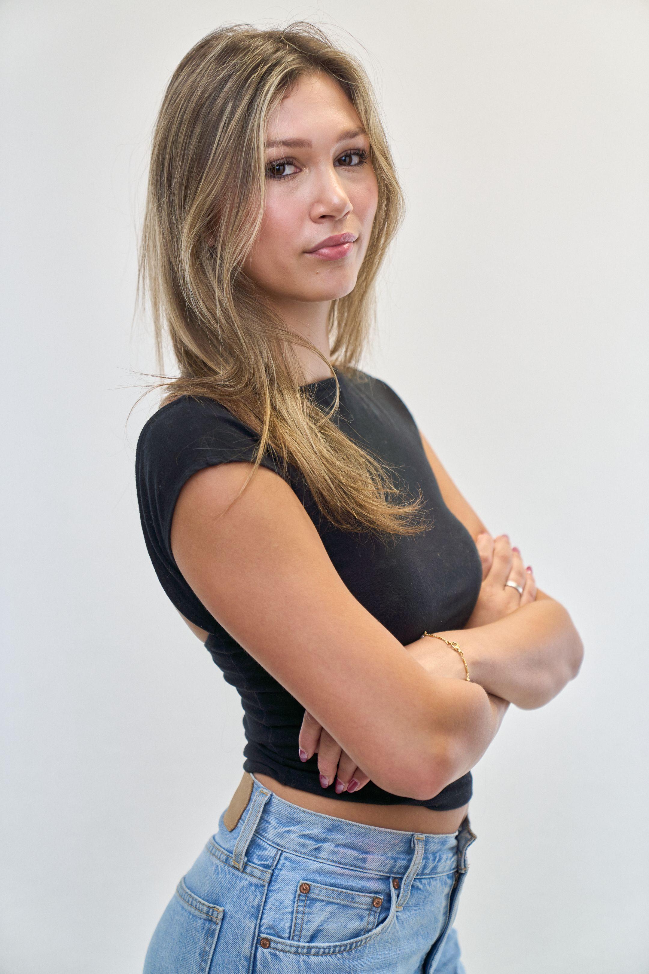 Model in a black cap-sleeve top and jeans turned to the side with arms folded — E-commerce & apparel photography, Austin Model in a black cap-sleeve top and jeans turned to the side with arms folded, against a white studio wall