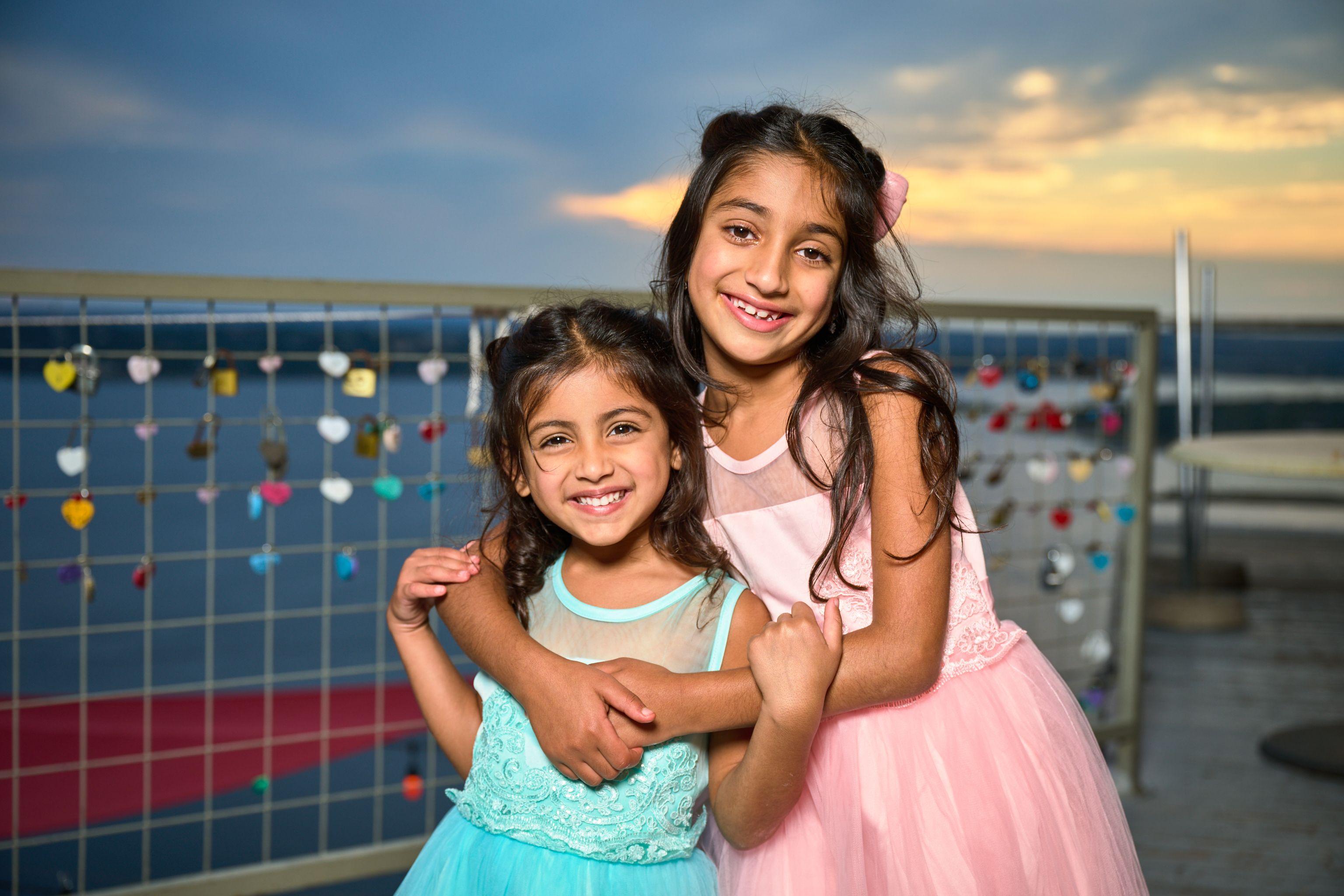 Two sisters in party dresses hugging on a deck at sunset, a lake and a fence of love locks — Mobile photo studio, Austin Two sisters in party dresses hugging on a deck at sunset, a lake and a fence of love locks behind them
