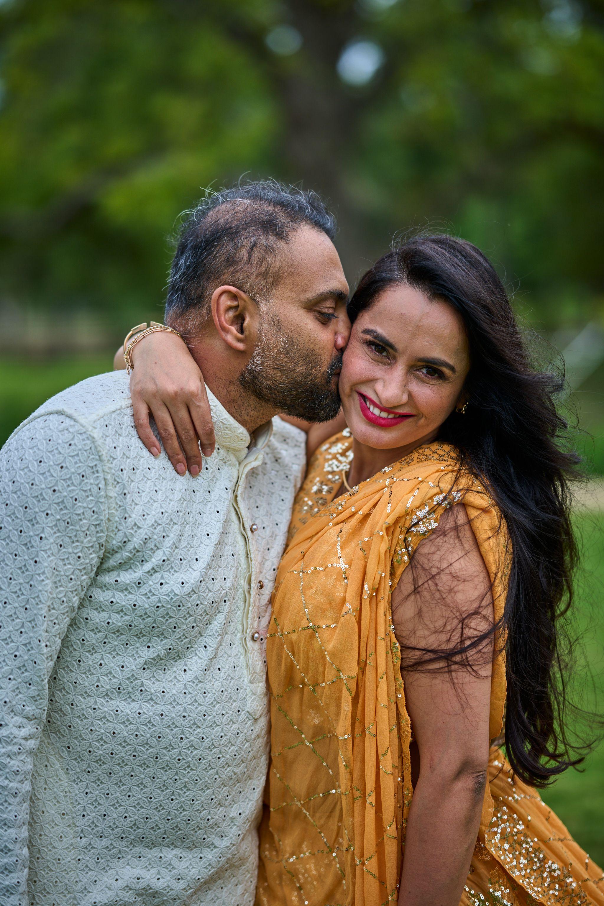 Husband in a pale kurta kissing his wife's cheek, she in a gold sari — Cedar Park Public Library photography, Austin A husband in a pale kurta kissing his wife's cheek, she in a gold sari.