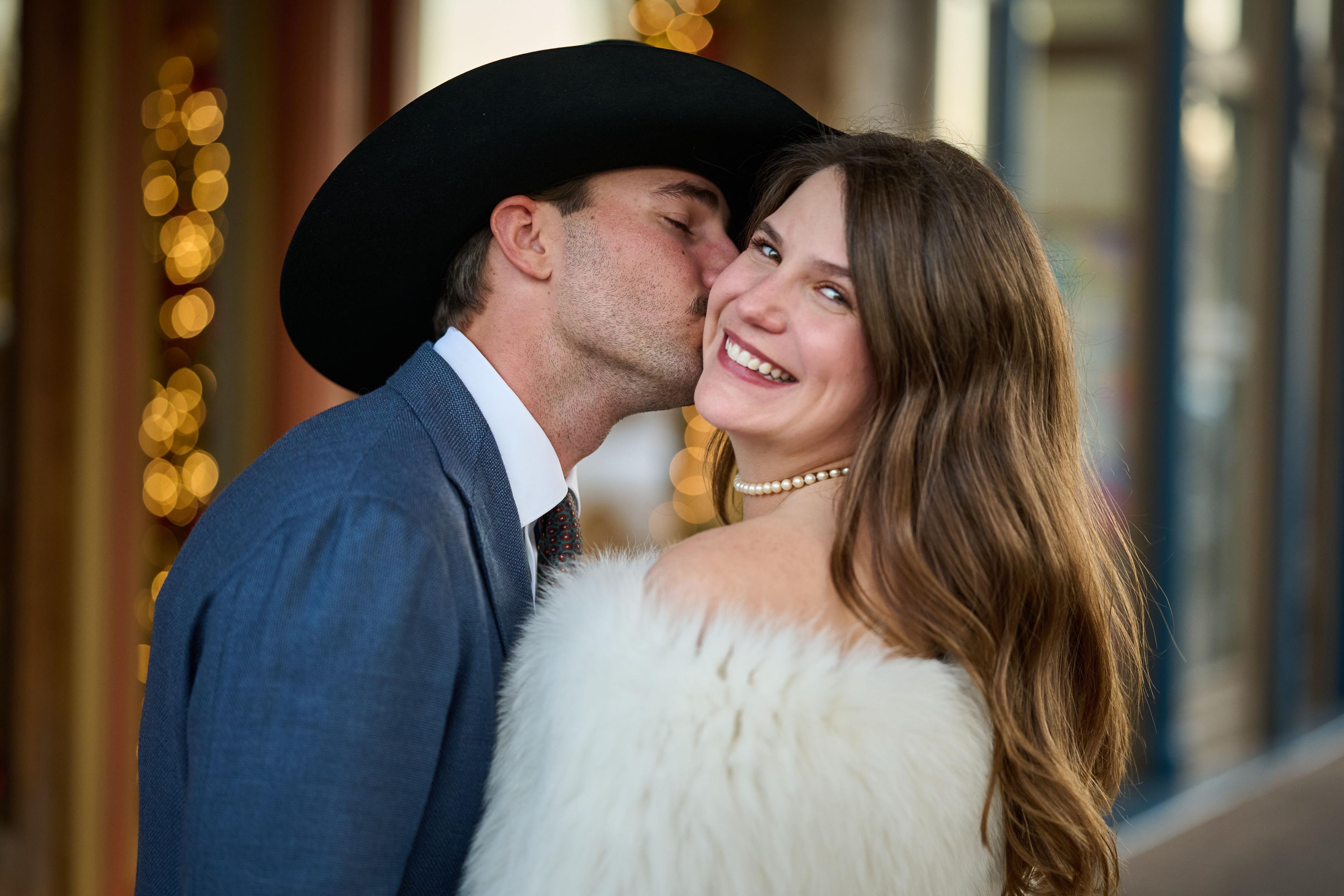 Groom in a cowboy hat kissing his bride's cheek as she laughs in a white fur wrap — Elopements photography, Austin A groom in a cowboy hat kissing his bride's cheek as she laughs in a white fur wrap.