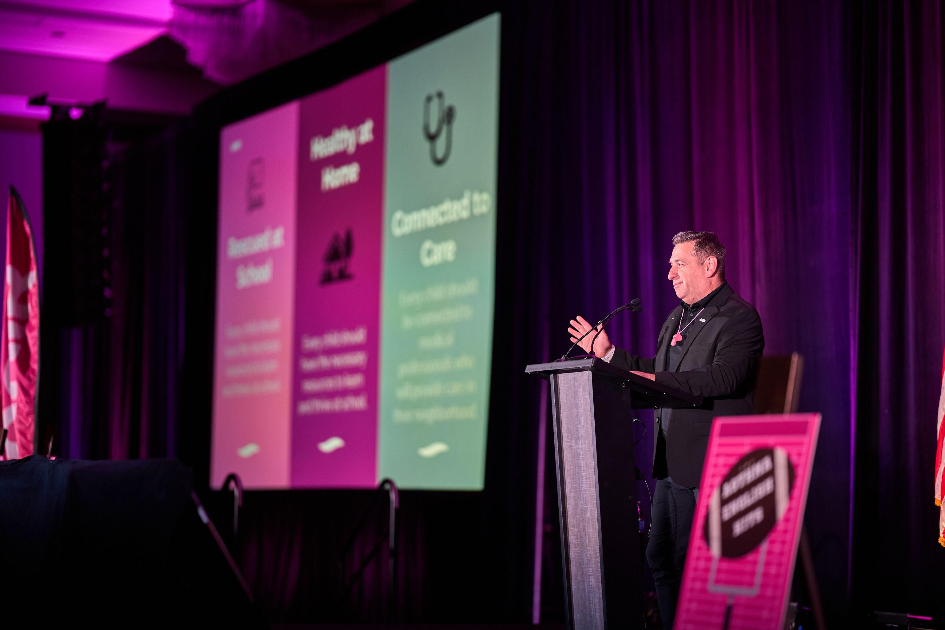 Speaker at a lectern beside a bright pink healthcare presentation slide — Conventions & Tradeshows photography, Austin A speaker at a lectern beside a bright pink healthcare presentation slide.