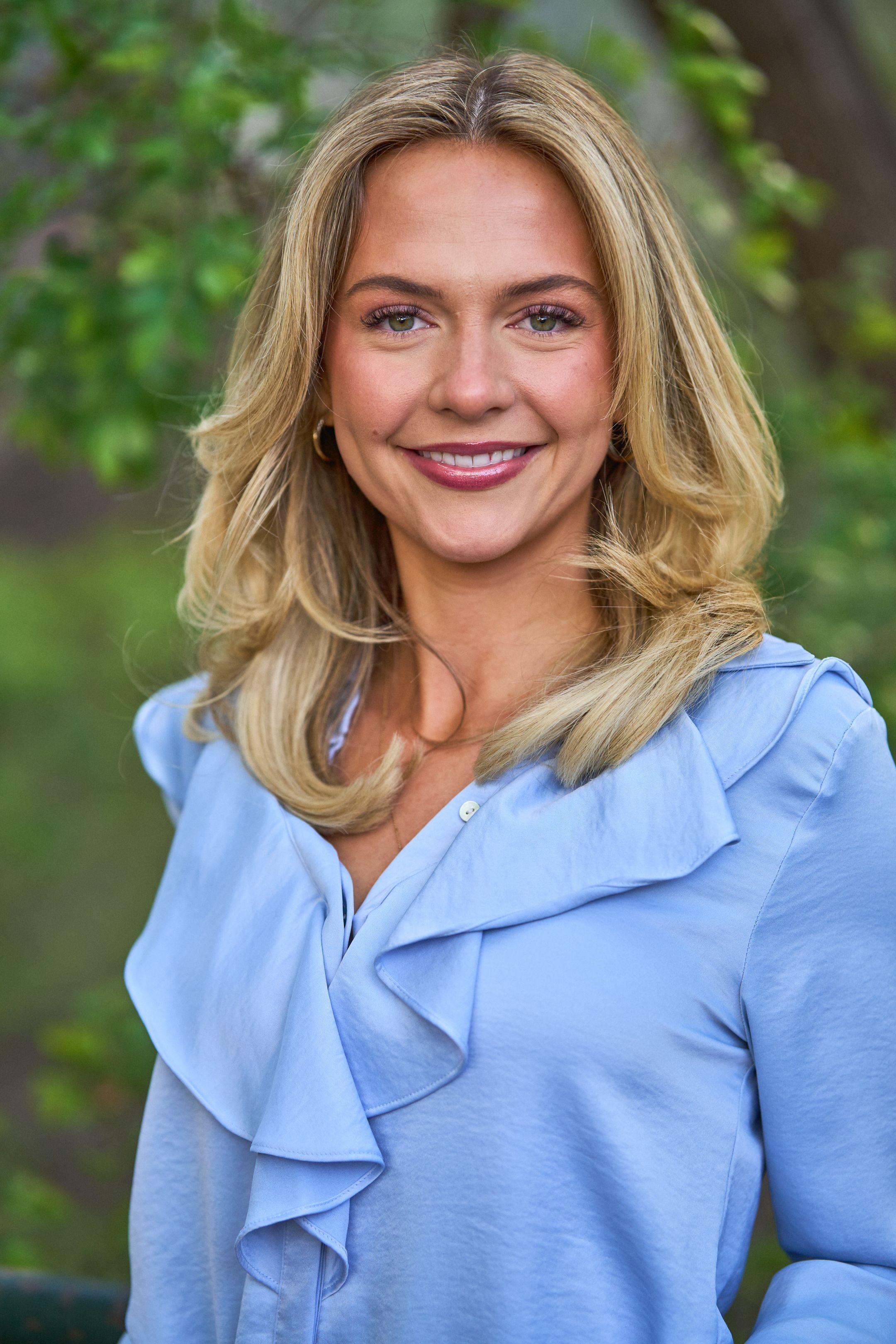 Woman in a pale blue ruffled blouse smiling, green foliage out of focus — Business headshots, Austin A woman in a pale blue ruffled blouse smiling, green foliage out of focus behind her