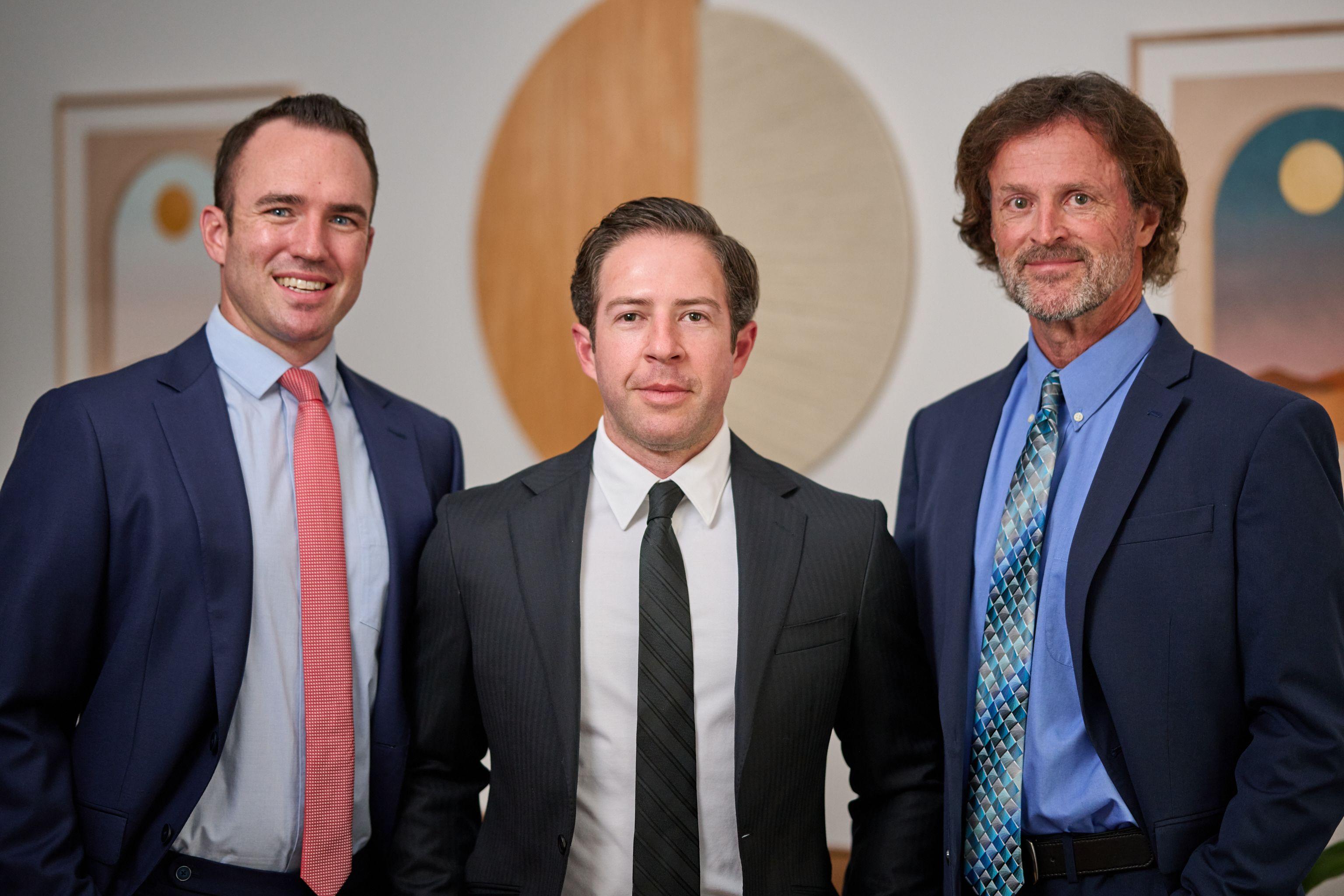 Three men in suits standing together in front of a curved wall panel in an office — Business headshots, Austin Three men in suits standing together in front of a curved wall panel in an office