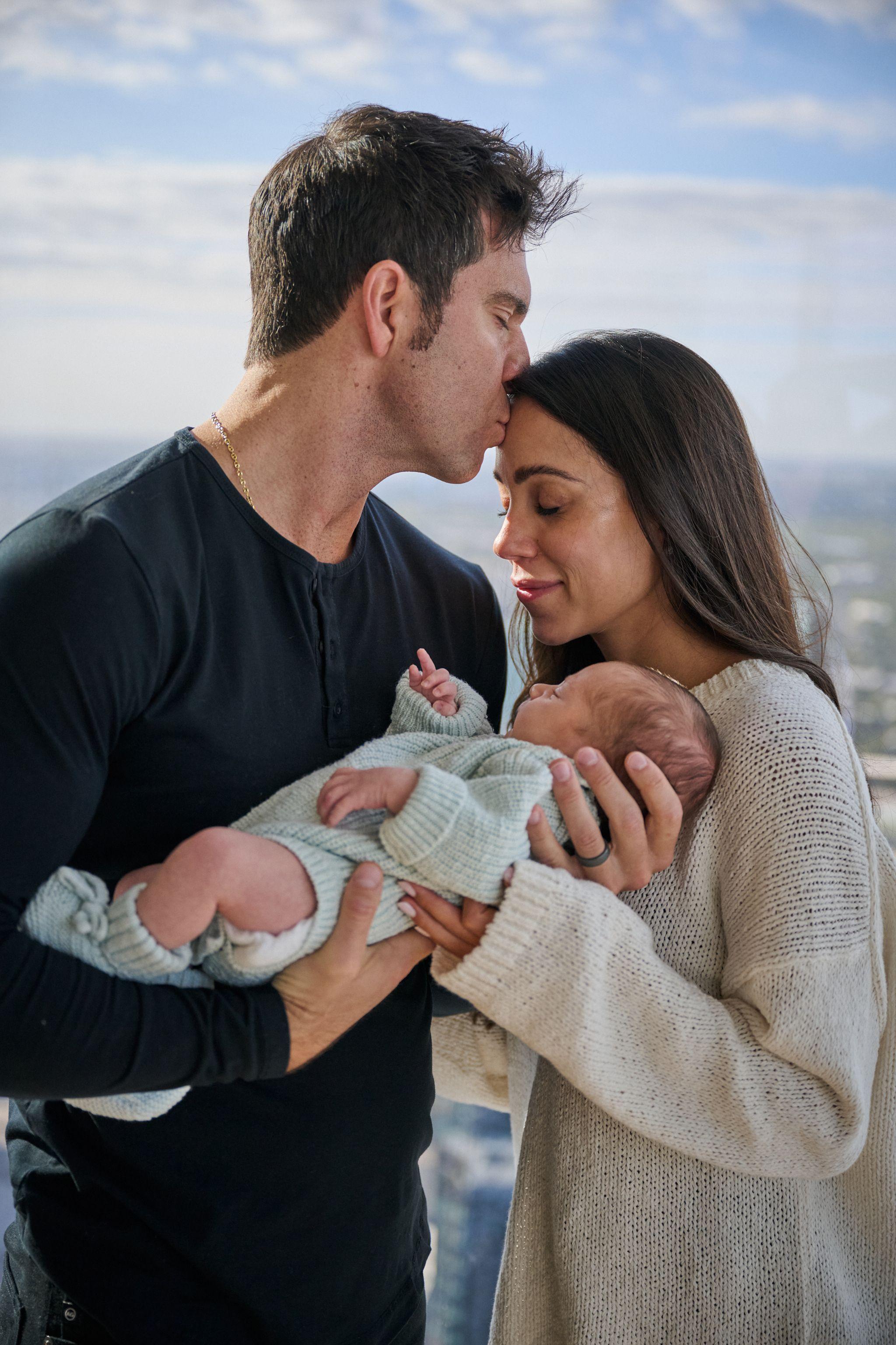 New parents standing together holding their swaddled newborn, city light behind them — Newborn photography, Austin New parents standing together holding their swaddled newborn, city light behind them.