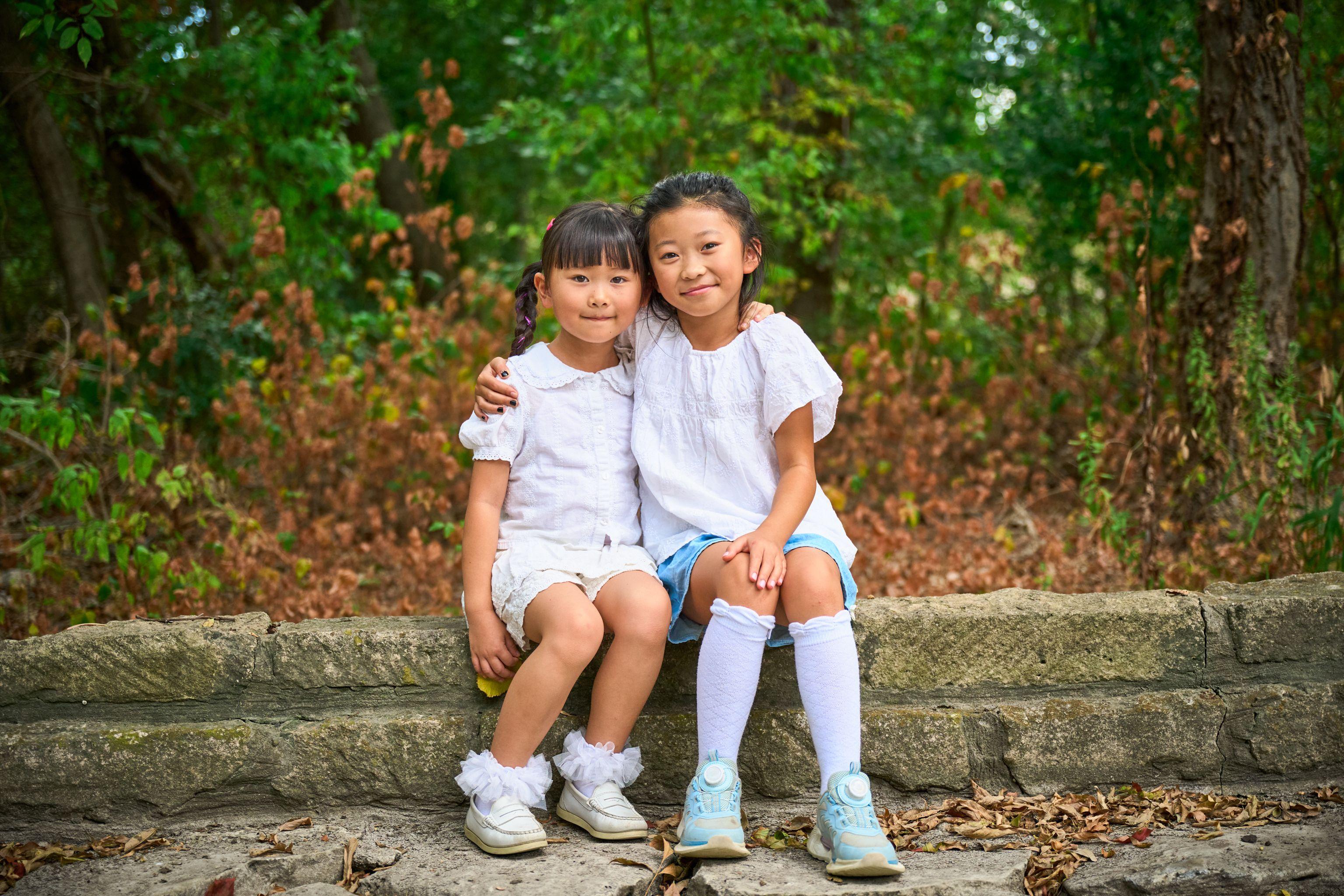 Two sisters in white sitting on a low stone wall, autumn leaves around them — Kids Portraits photography, Austin Two sisters in white sitting on a low stone wall, autumn leaves around them.