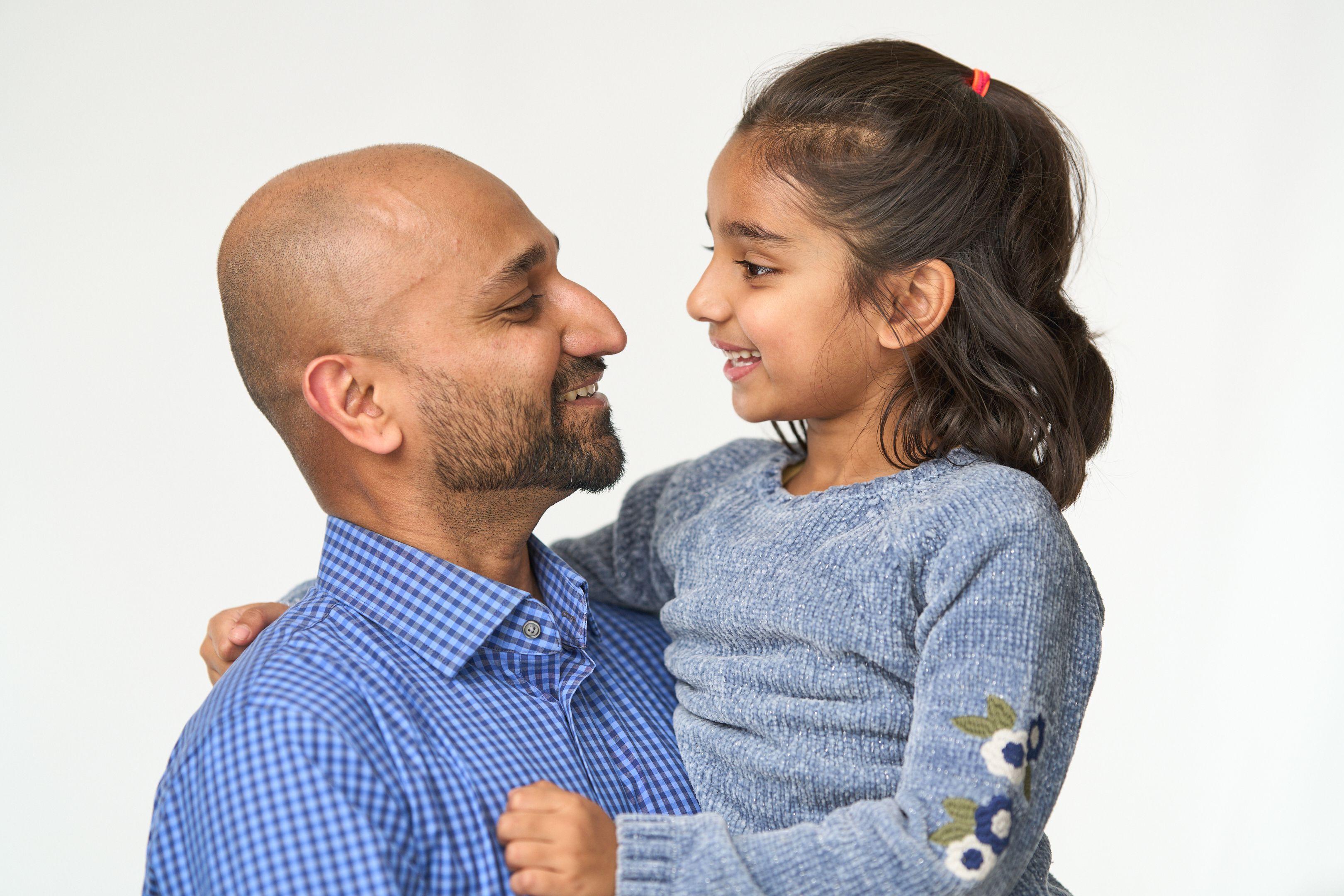 Bald man in a blue checked shirt holding his daughter, the two of them looking at each other — Commercial photo studio, Austin Bald man in a blue checked shirt holding his daughter, the two of them looking at each other on white seamless