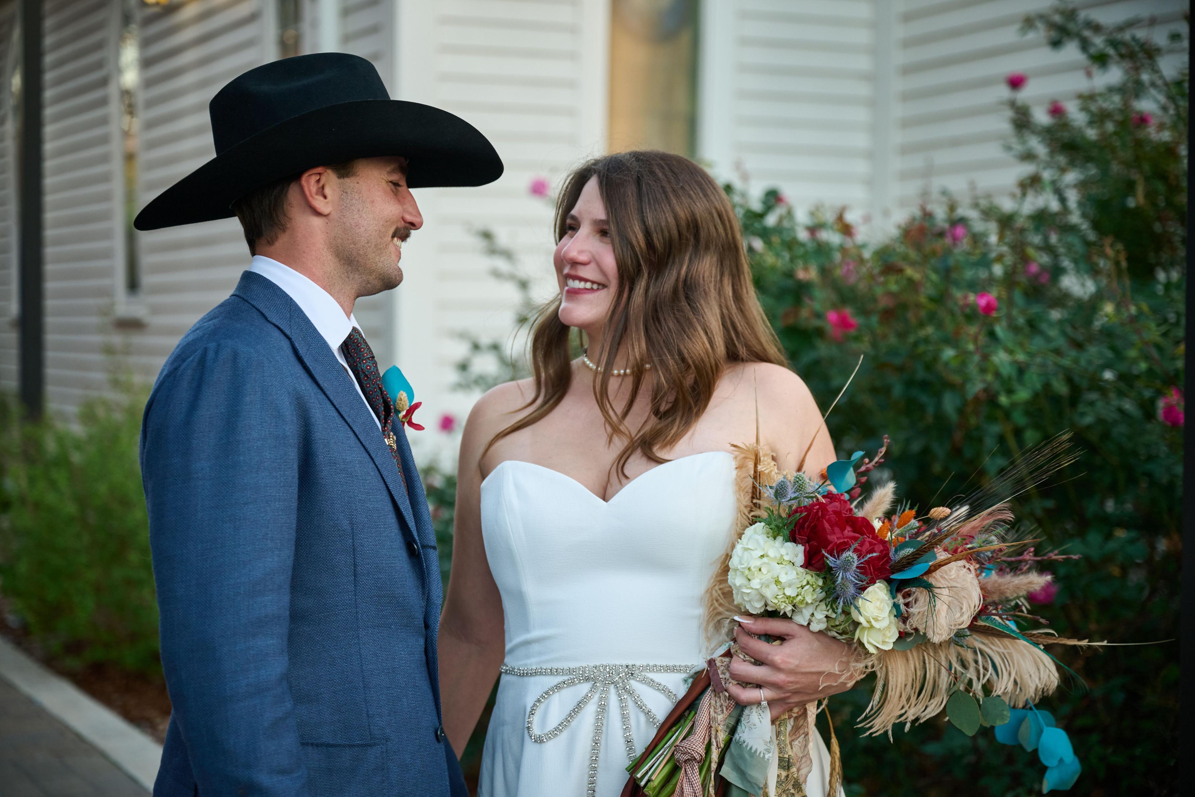 Bride and groom standing together beside a rose bush, she in white holding a red — Elopements photography, Austin A bride and groom standing together beside a rose bush, she in white holding a red and teal bouquet.