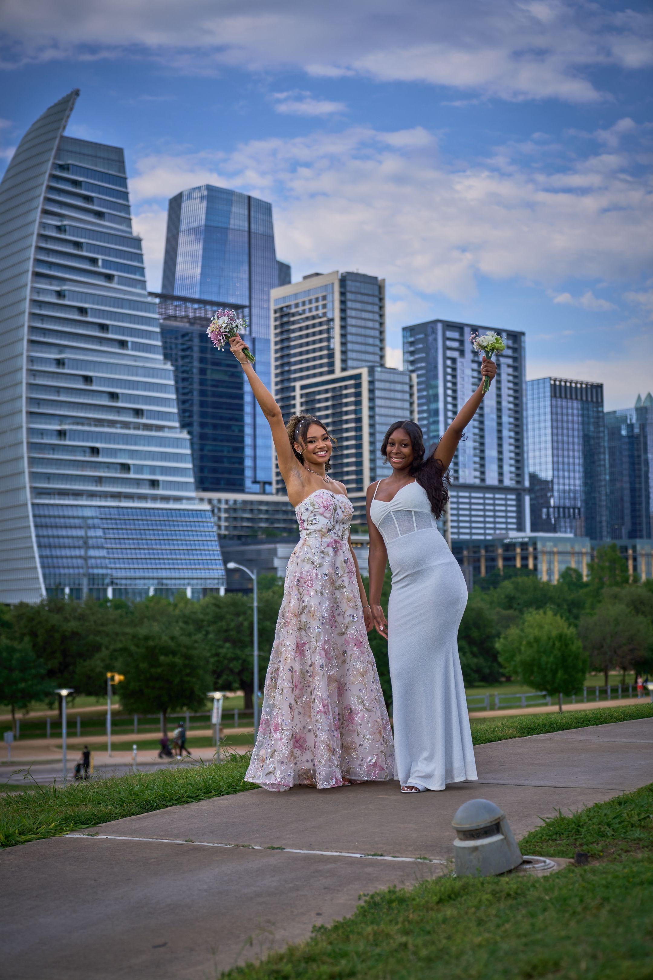 Two women in formal gowns raising their arms on a park path with the Austin skyline — Butler Metro Park photography, Austin Two women in formal gowns raising their arms on a park path with the Austin skyline behind.